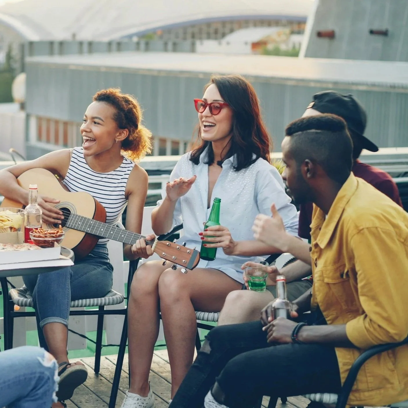 Group of friends sitting on a rooftop, enjoying music, drinks, and conversation, with some playing guitar and others sharing smiles.