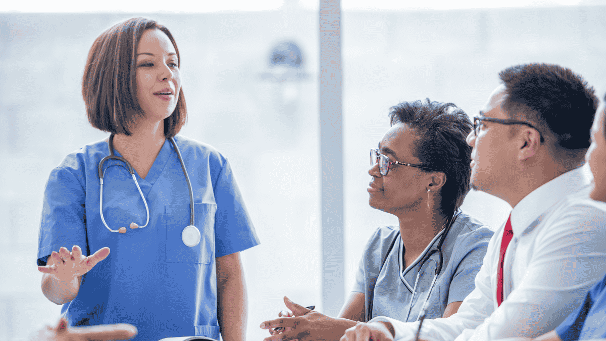 Female nurse or doctor in blue scrubs speaking to a group of healthcare professionals in an office setting.