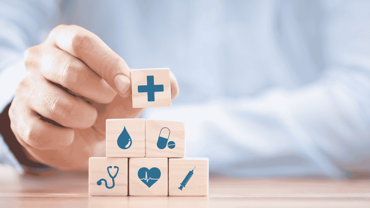 Hand stacking wooden blocks with healthcare symbols, including a medical cross, blood drop, pill, stethoscope, heartbeat, and syringe.