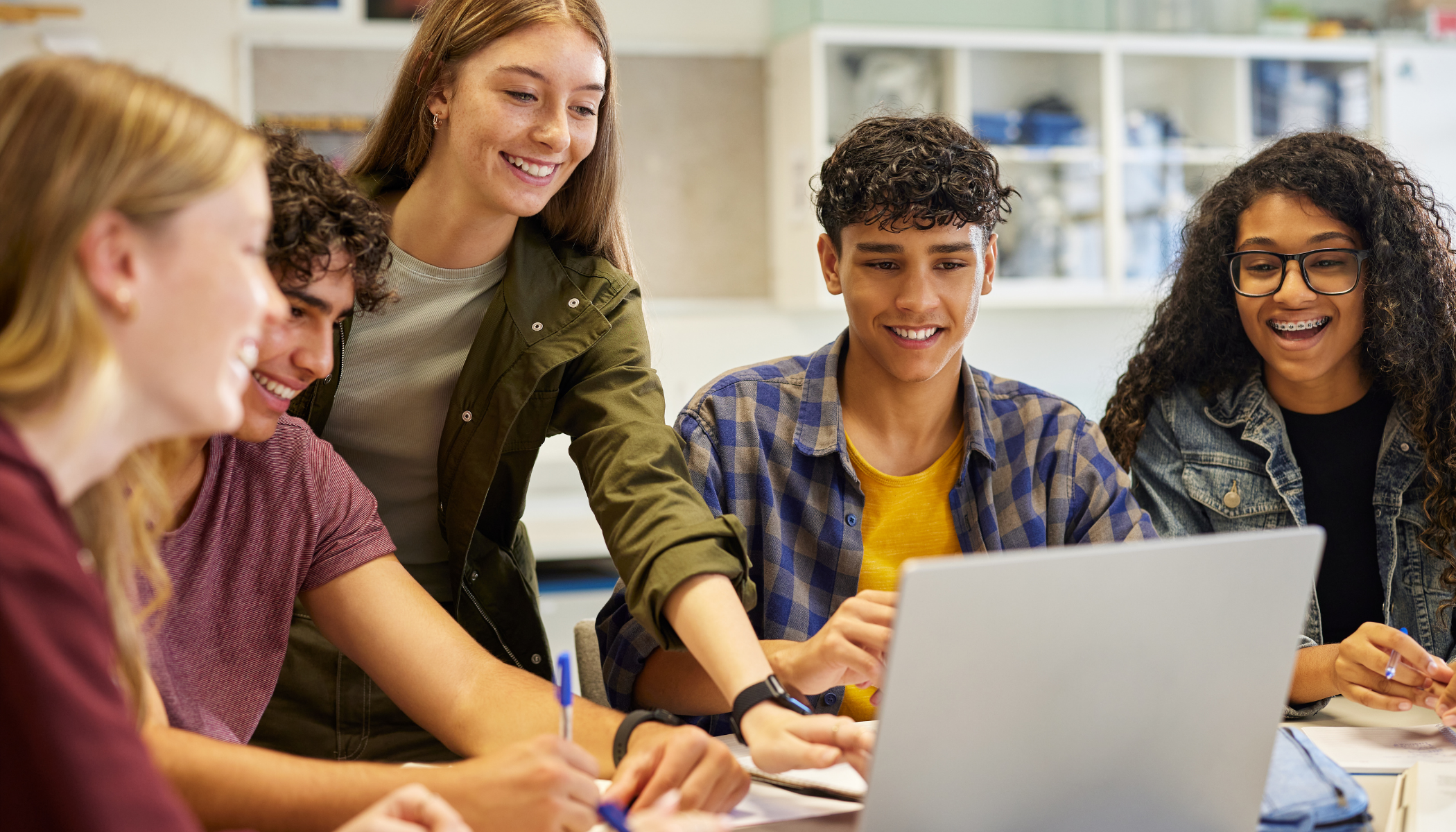 Group of diverse teenagers gathered around a laptop, smiling and collaborating in a classroom.