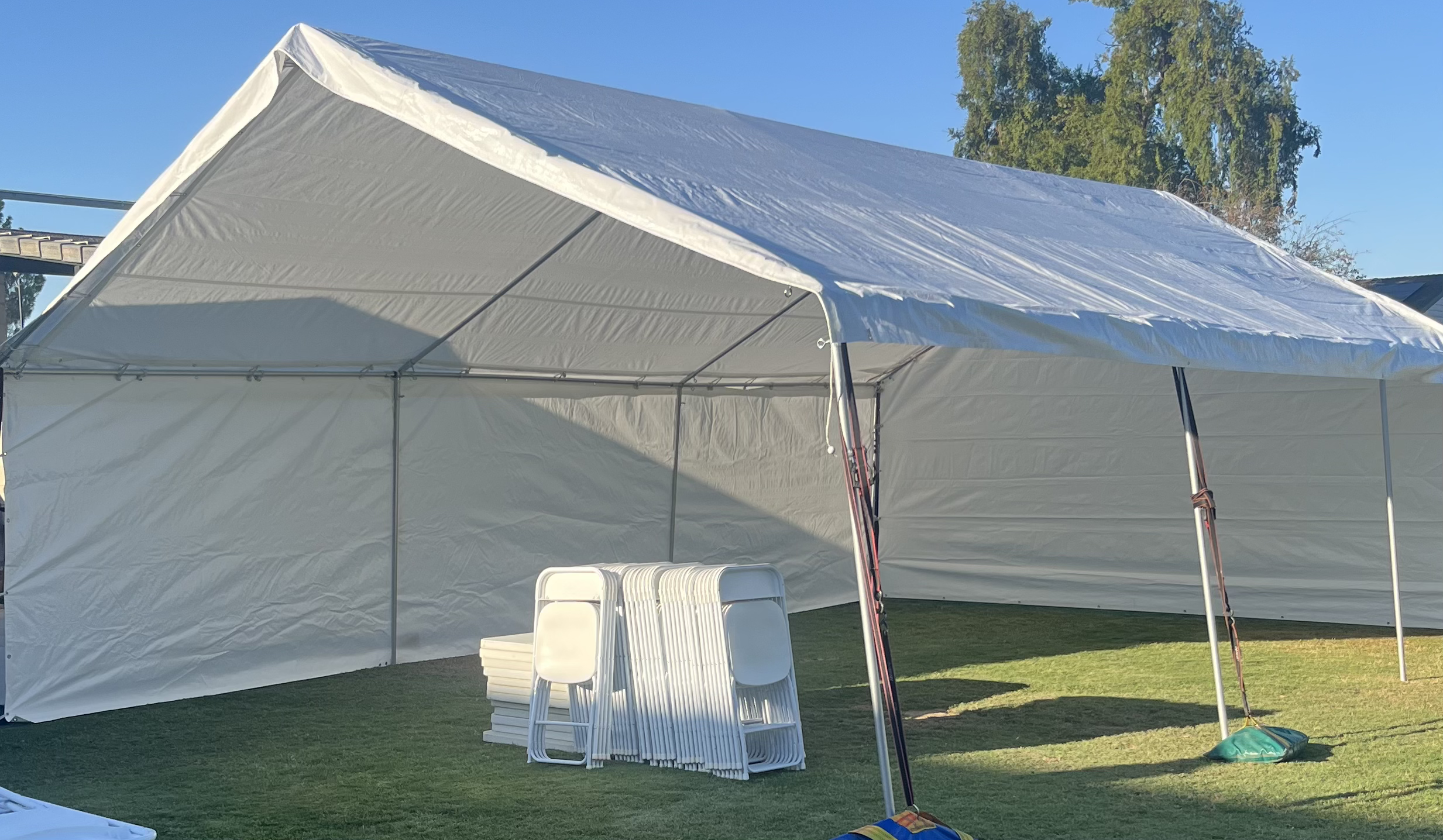 Empty white event tent with stacked chairs and a pool cleaning net on a grassy lawn, with trees and a clear blue sky in the background.