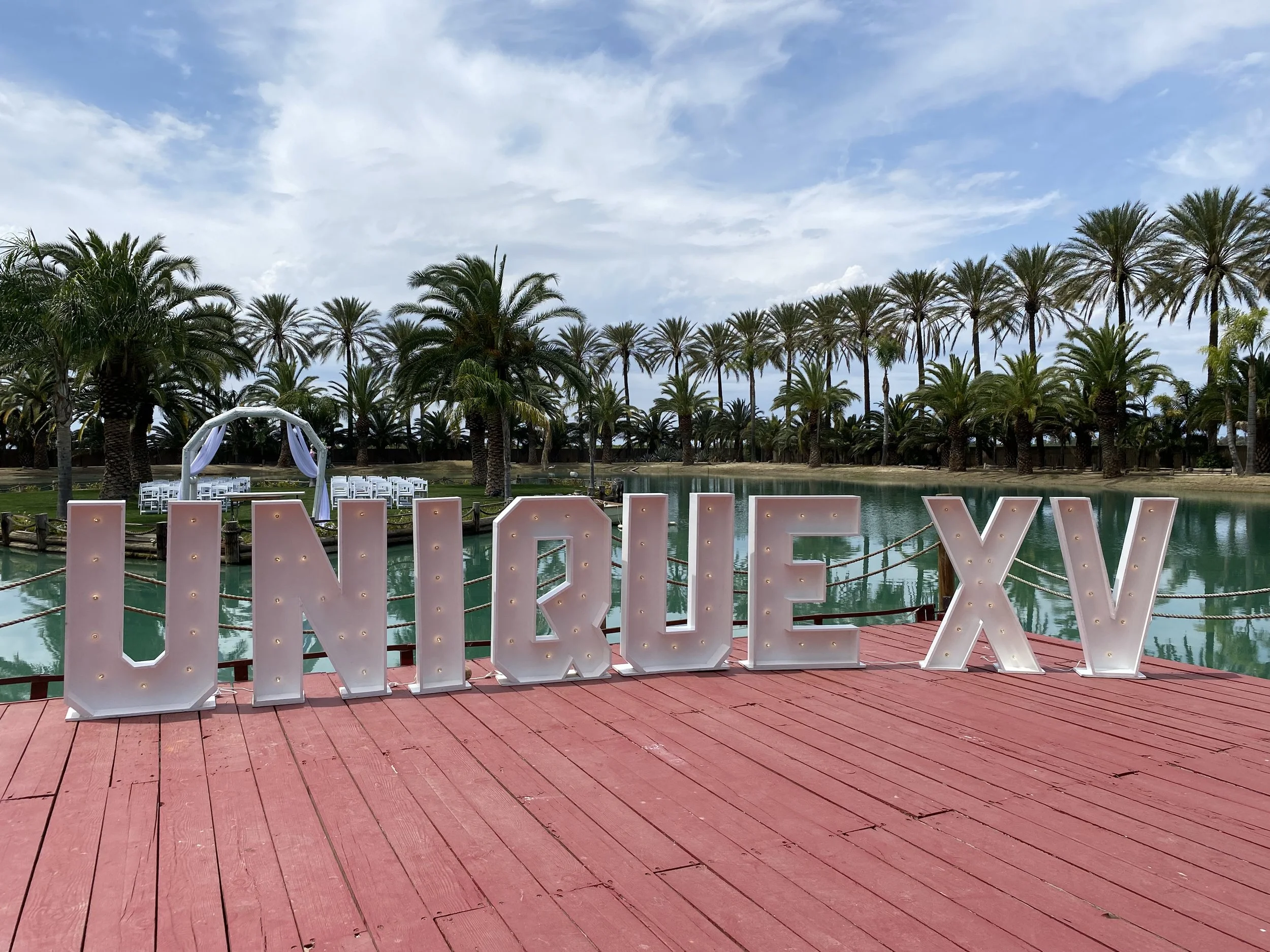 Large illuminated letters spelling 'UNIRIO XV' on a red wooden deck near a water body, with palm trees and chairs in the background under a cloudy sky.