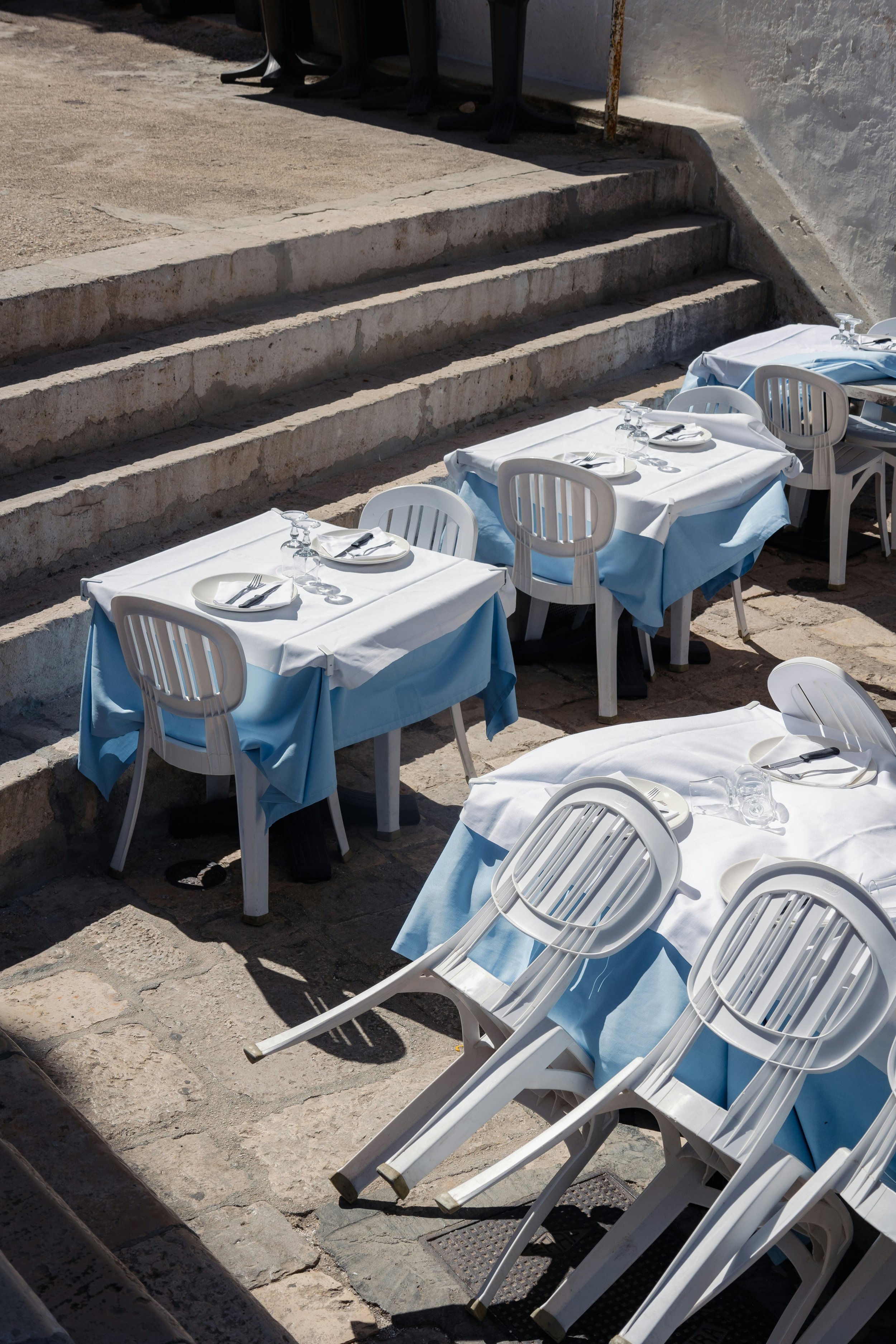 Empty outdoor restaurant tables with white and light blue tablecloths, set with plates, silverware, and wine glasses, some chairs are overturned on the stone floor, and a staircase is visible in the background.