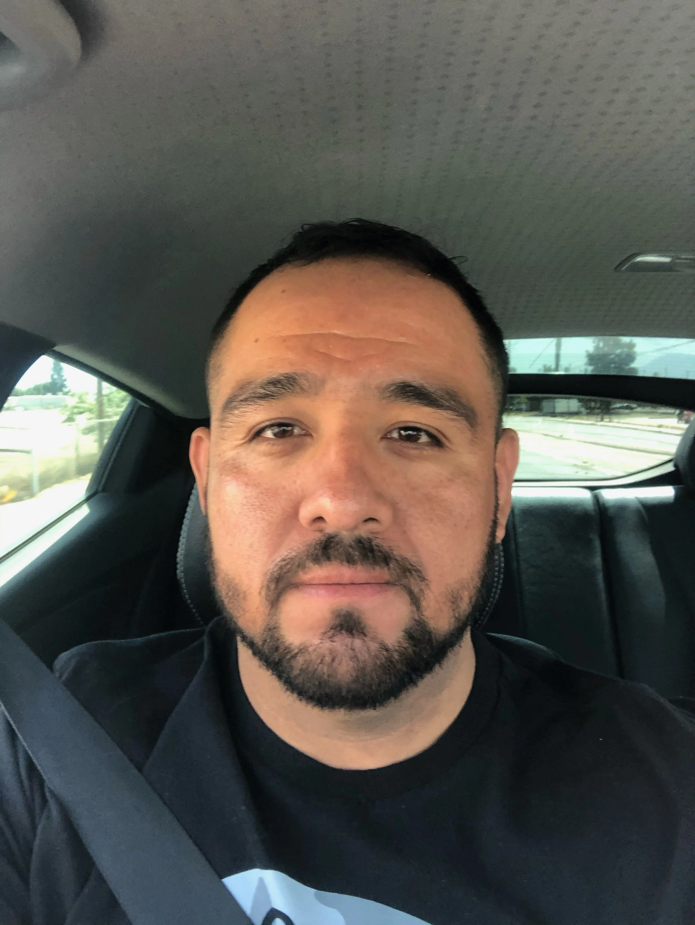 A man with short black hair, a beard, and mustache sitting in the back seat of a car, wearing a black T-shirt, with a serious expression, and the car's interior, windows, and outside scenery are visible.