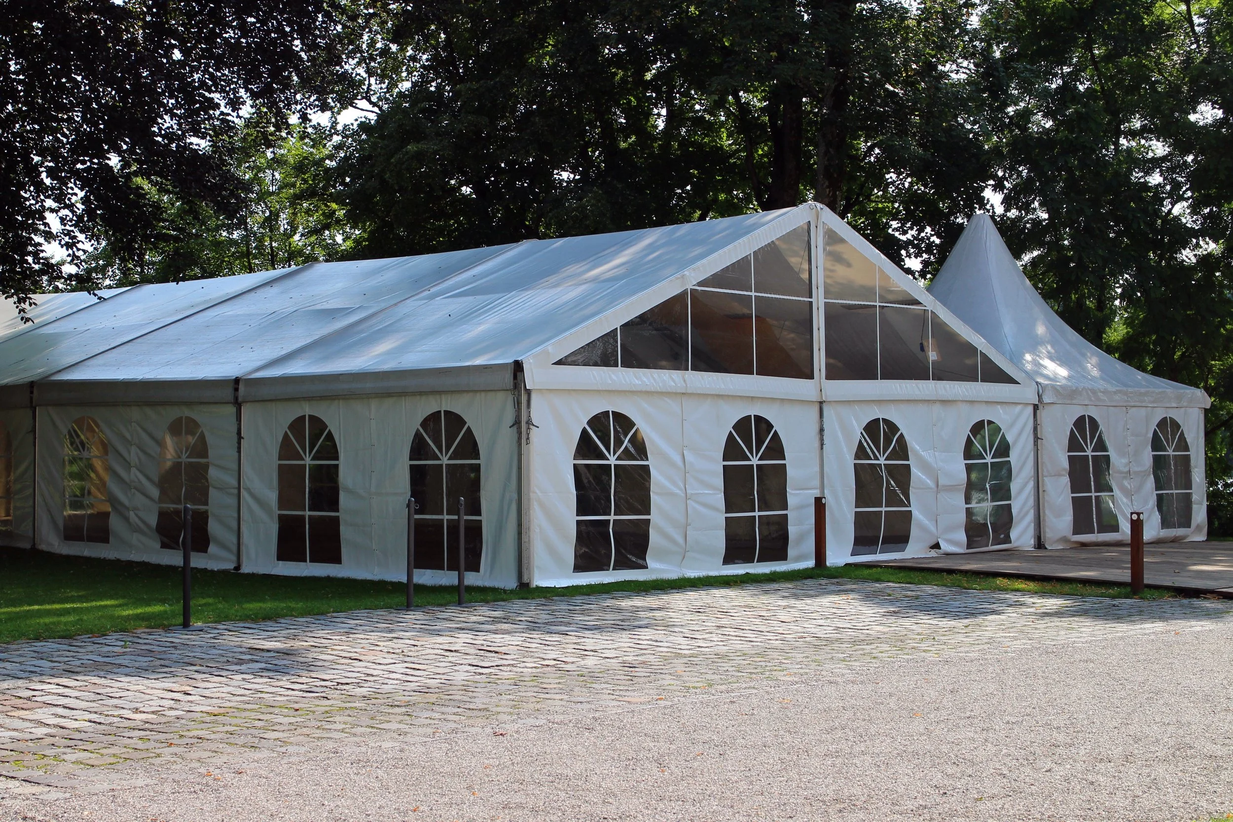 Large white event tent with arched windows and a peaked roof, set up outdoors on a cobblestone and grass area, surrounded by trees.