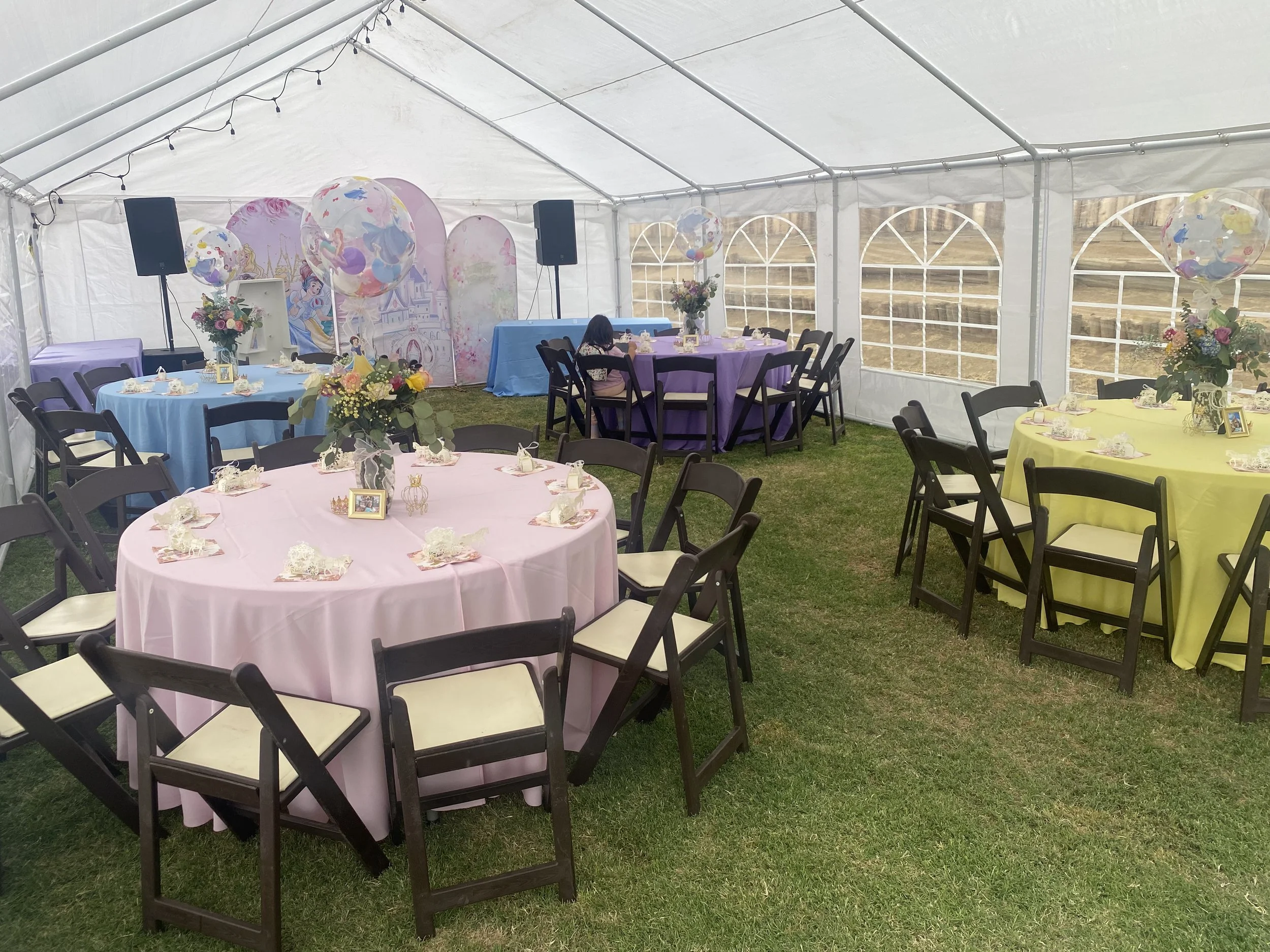 Decorated party tent with pastel-colored round tables, floral centerpieces, and helium balloons, set up for a celebration with chairs arranged around the tables.