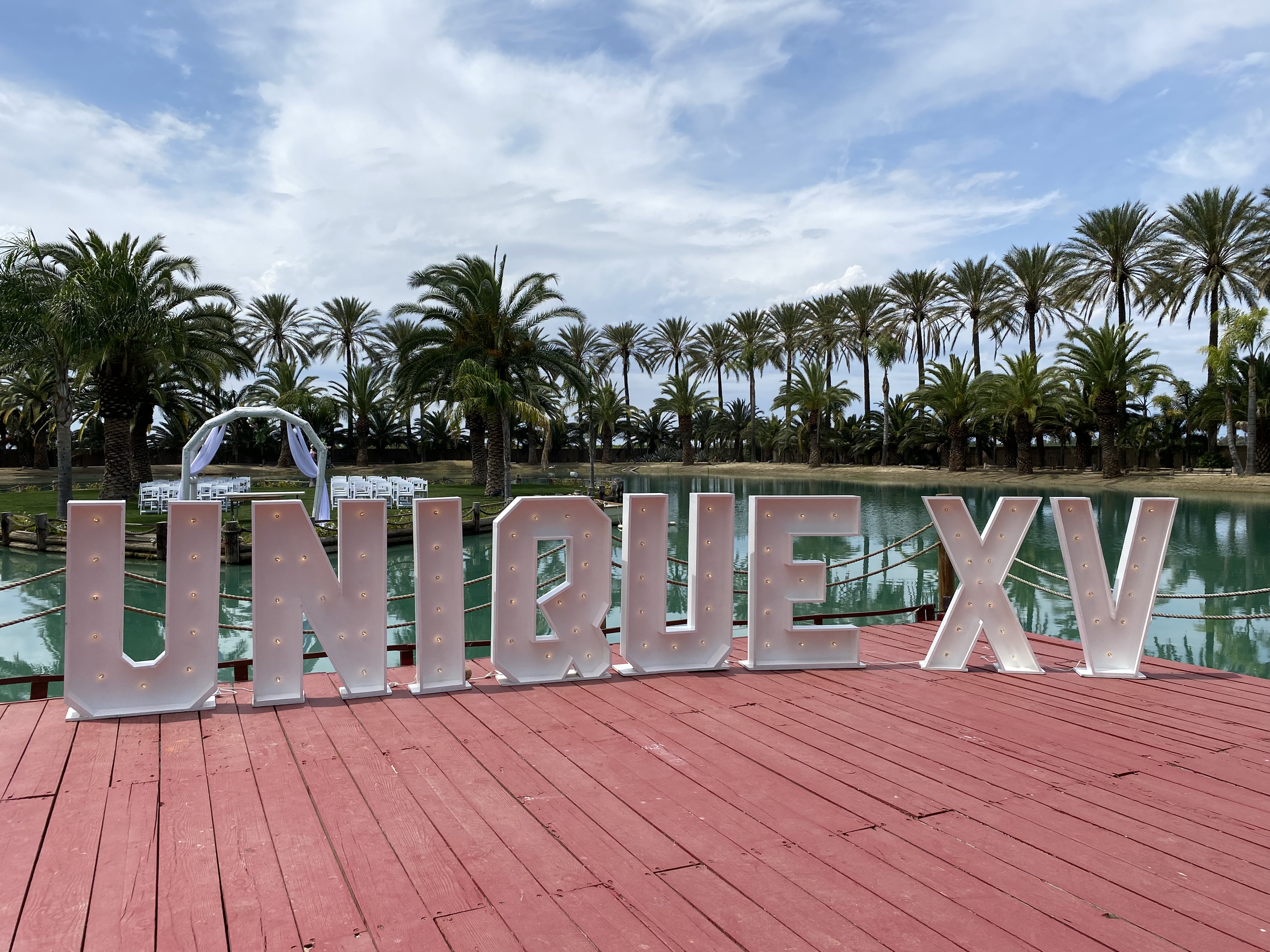 Large decorative letters spelling "UNIRIE XV" on a red wooden pier by a lagoon, with palm trees and a wedding arch in the background, under a partly cloudy sky.