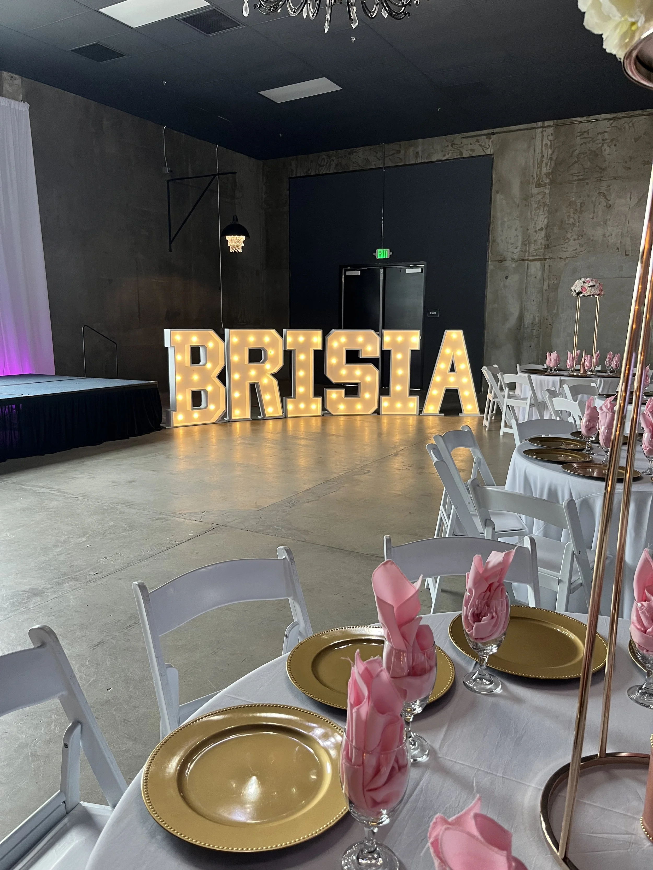 Event hall with large illuminated sign spelling 'BRISIA' in front of the stage, decorated round tables with white tablecloths, gold plates, pink napkins in glasses, chairs, and elegant decor.