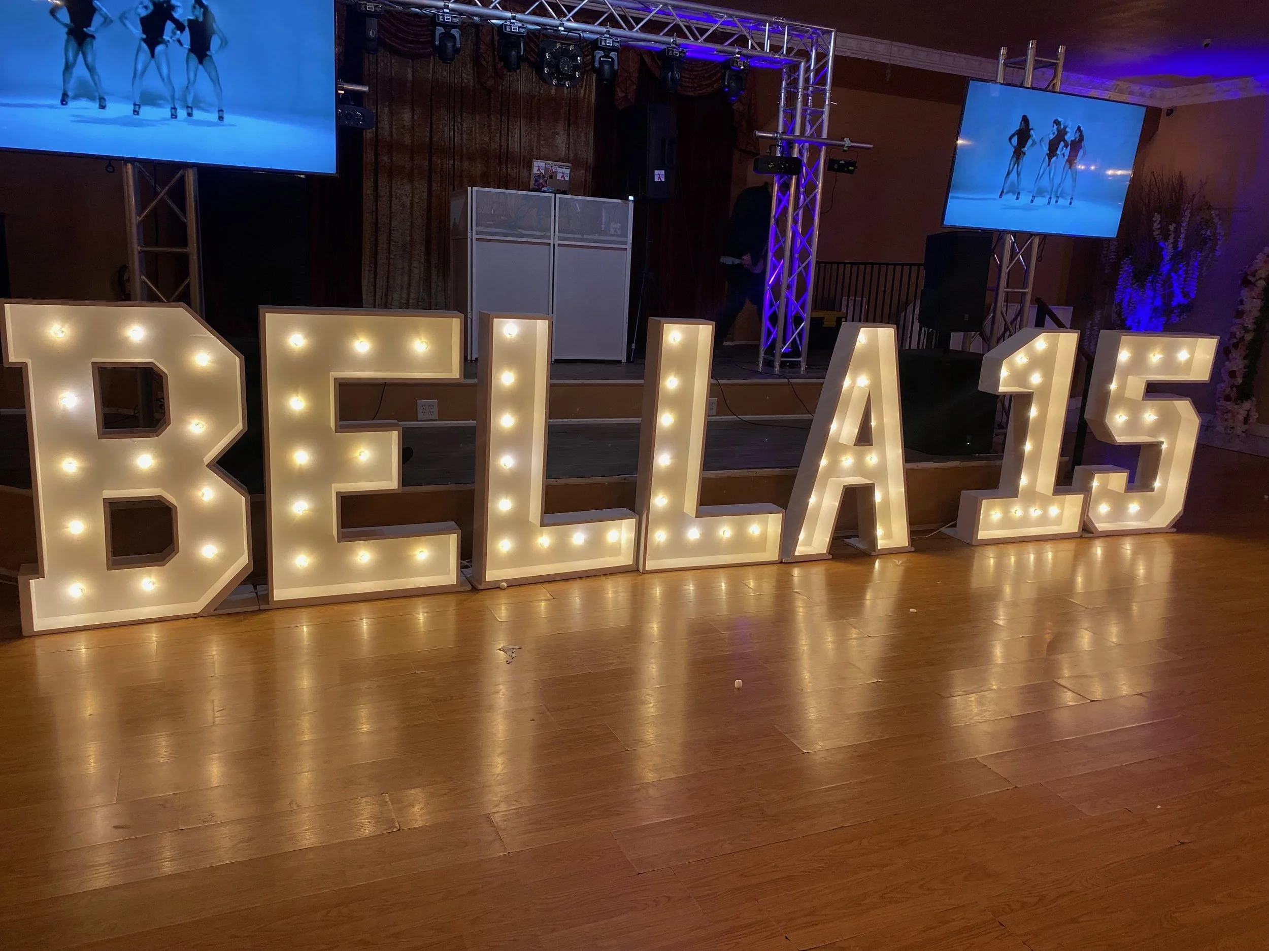 Large illuminated sign spelling out 'BELLA 15' with stage and video screens in the background at an event or celebration.