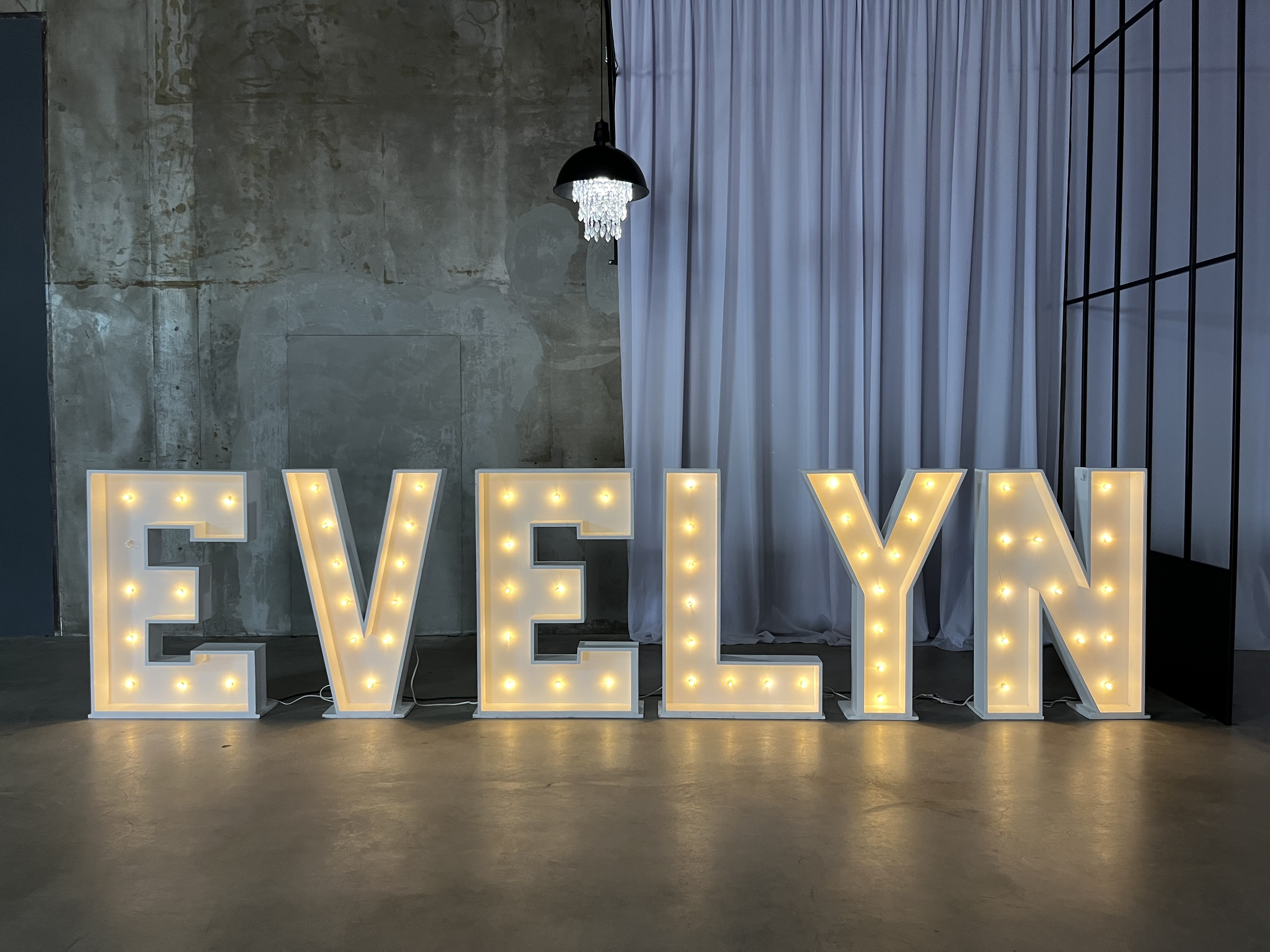 Large illuminated marquee letters spelling 'EVELYN' on a polished floor against an industrial concrete wall and white curtain backdrop.