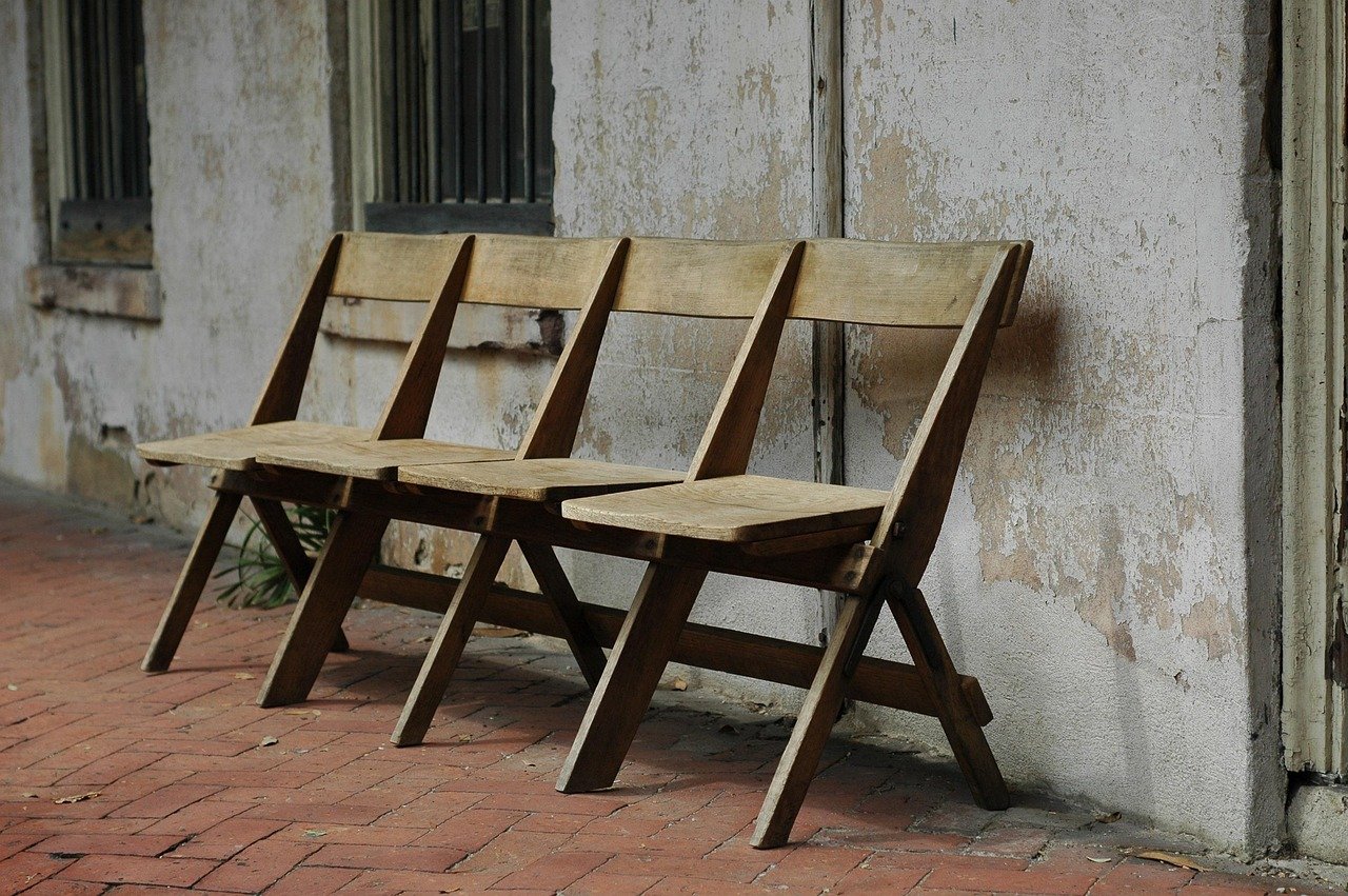 Three wooden chairs are lined up outside against a weathered, white-painted wall with two windows. The chairs are unoccupied and placed on a brick sidewalk.
