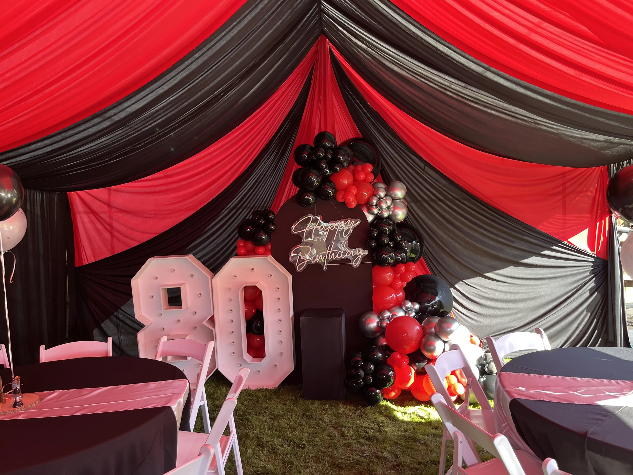 Birthday celebration setup with large illuminated number 80, black, red, and silver balloon decorations, pink and black tablecloths, and black, red, and silver balloons on a backdrop with black and red drapes.