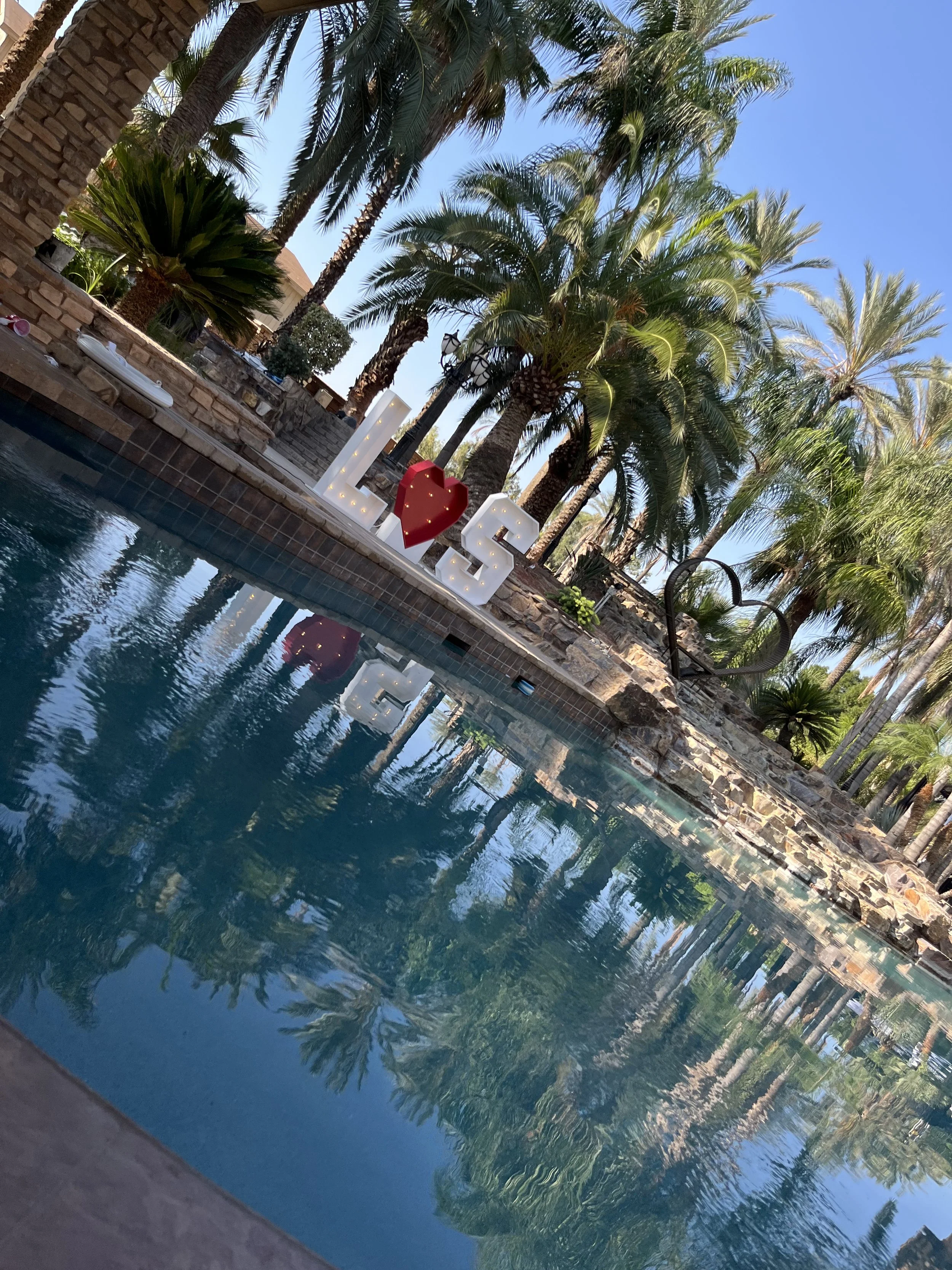 Sunset view of a backyard swimming pool surrounded by palm trees and decorative stones, with large letter signs spelling 'LOVE' and a red heart.