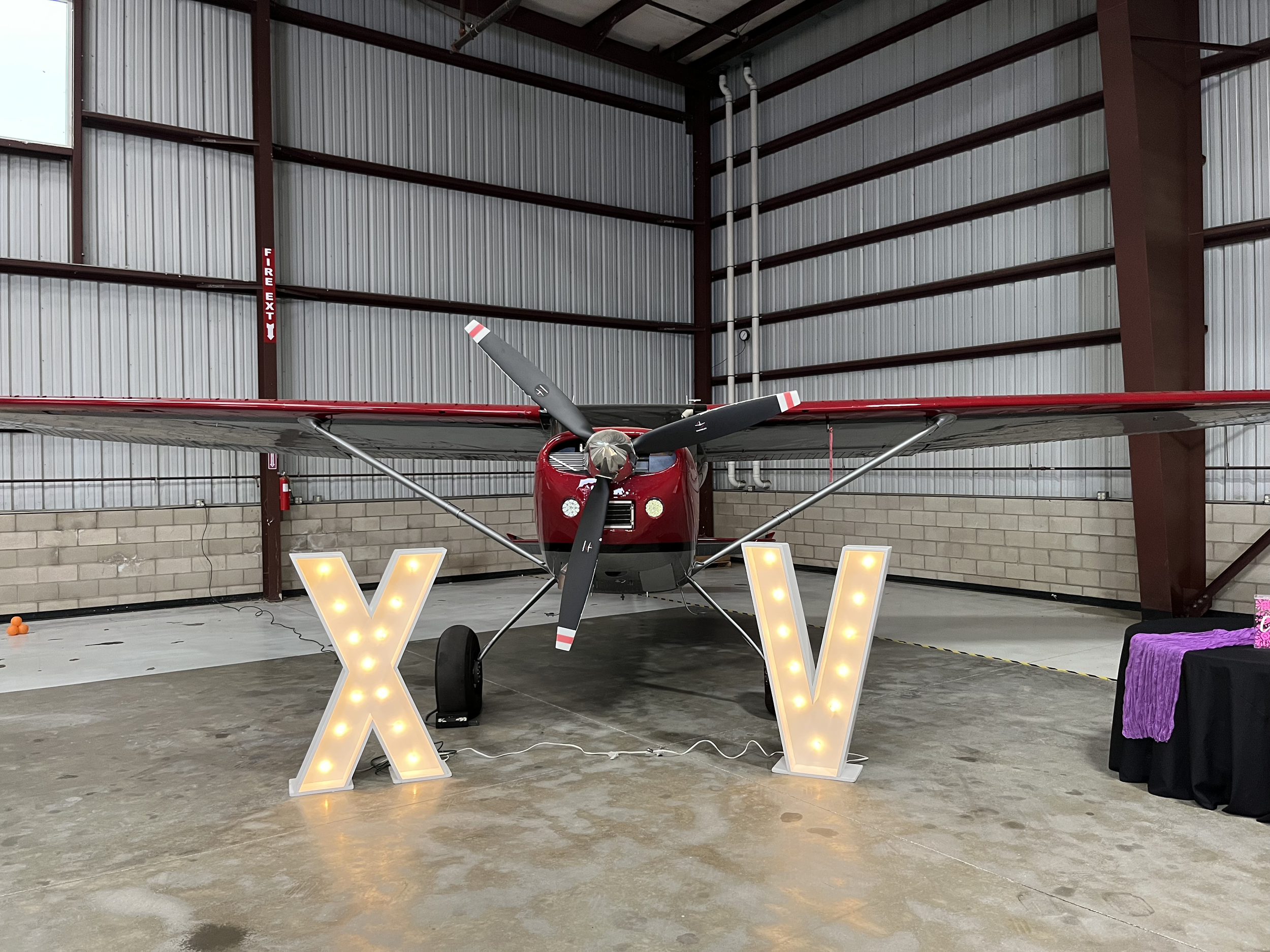 A small red and black airplane inside a hangar, decorated with large illuminated letters "X" and "V" in front of it.