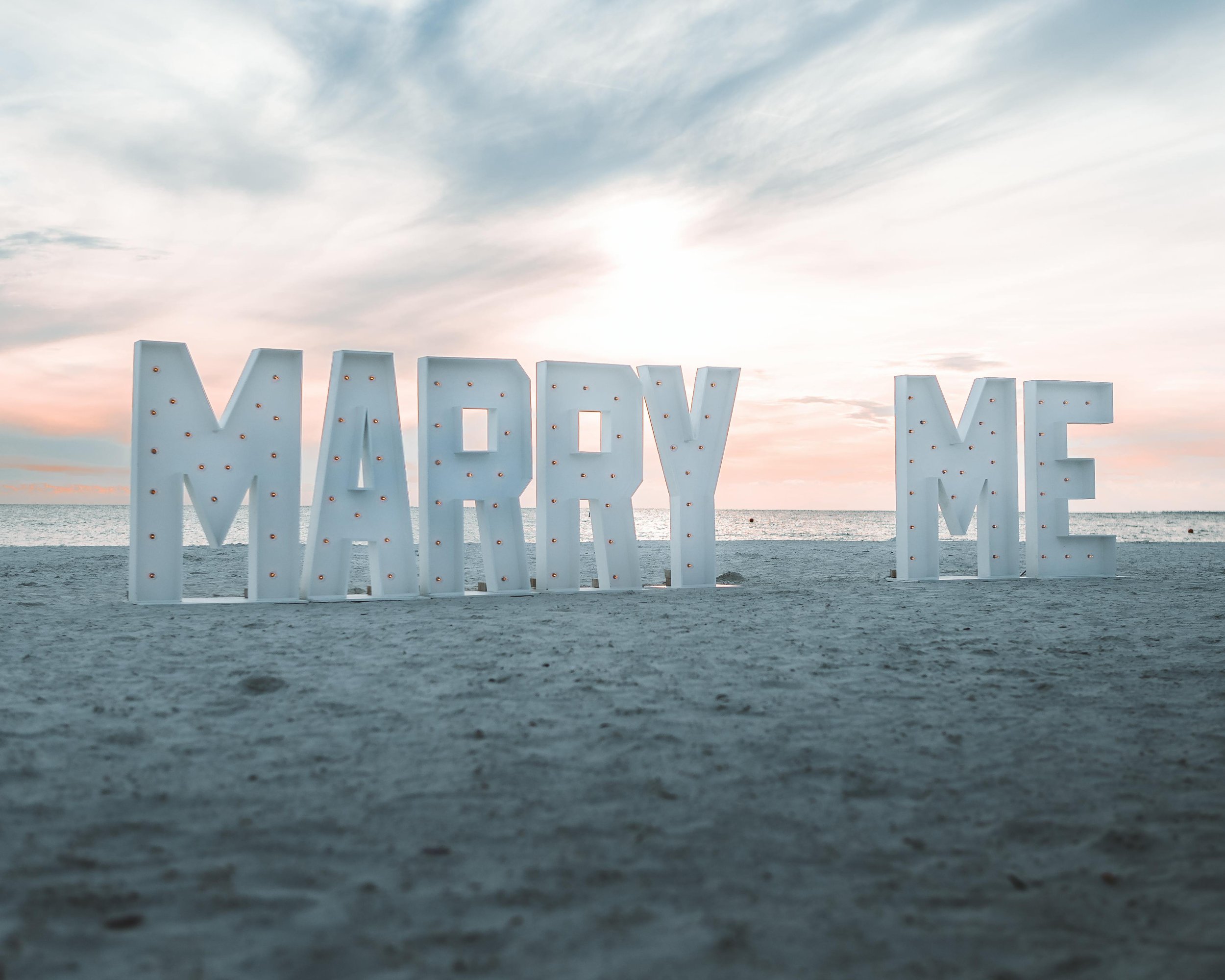 Large white illuminated letters spelling 'MARRY ME' on a beach at sunset.
