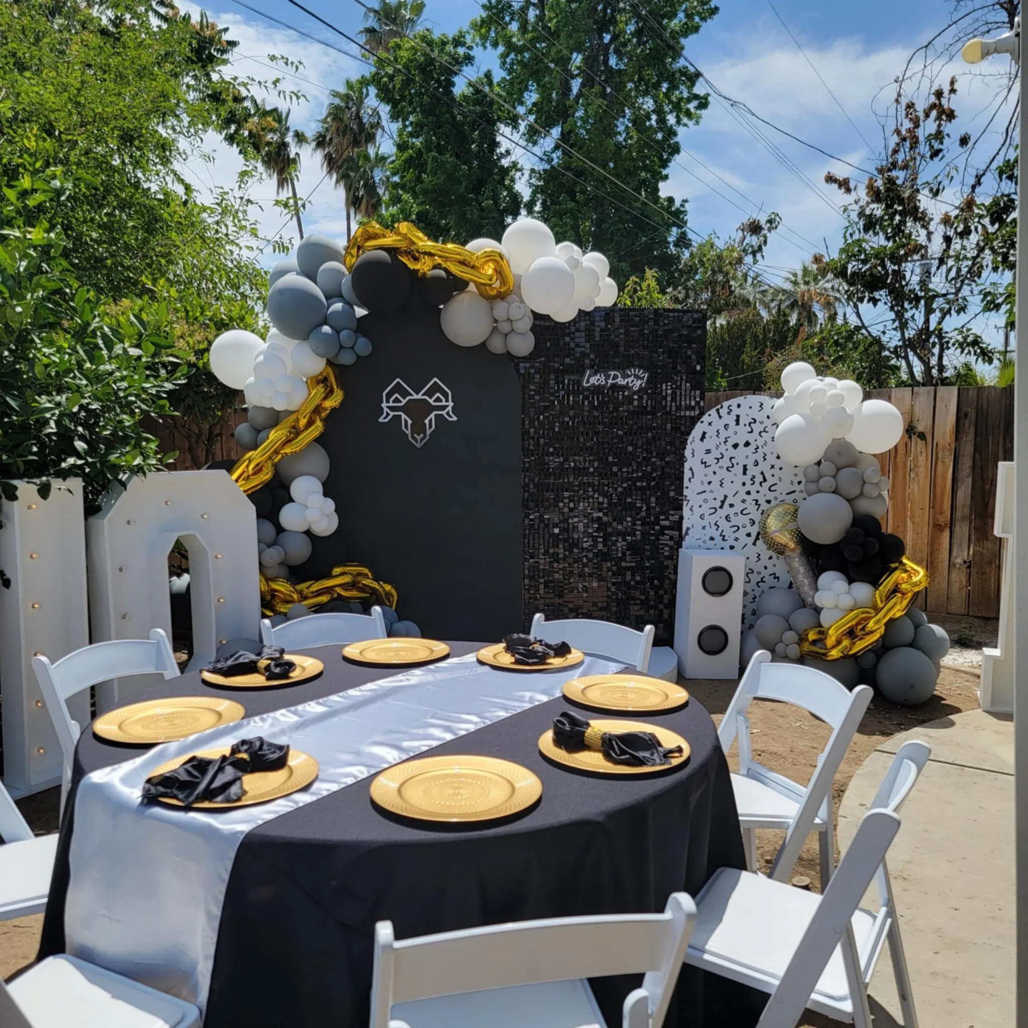 Outdoor party setup with a round table covered in black and white tablecloth, gold plates, black napkins, and white chairs, in front of a black backdrop decorated with balloons in black, white, gray, and gold, and a black and white patterned panel, surrounded by greenery and a wooden fence under a partly cloudy sky.