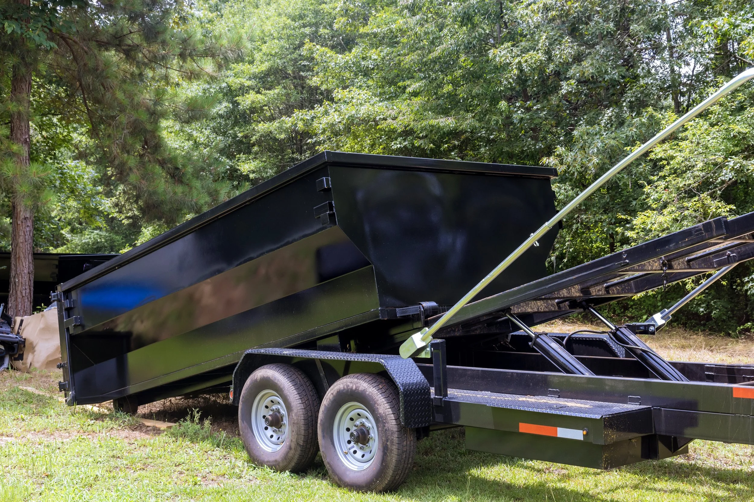 A black trailer with a hydraulic lift system on grass, surrounded by green trees.