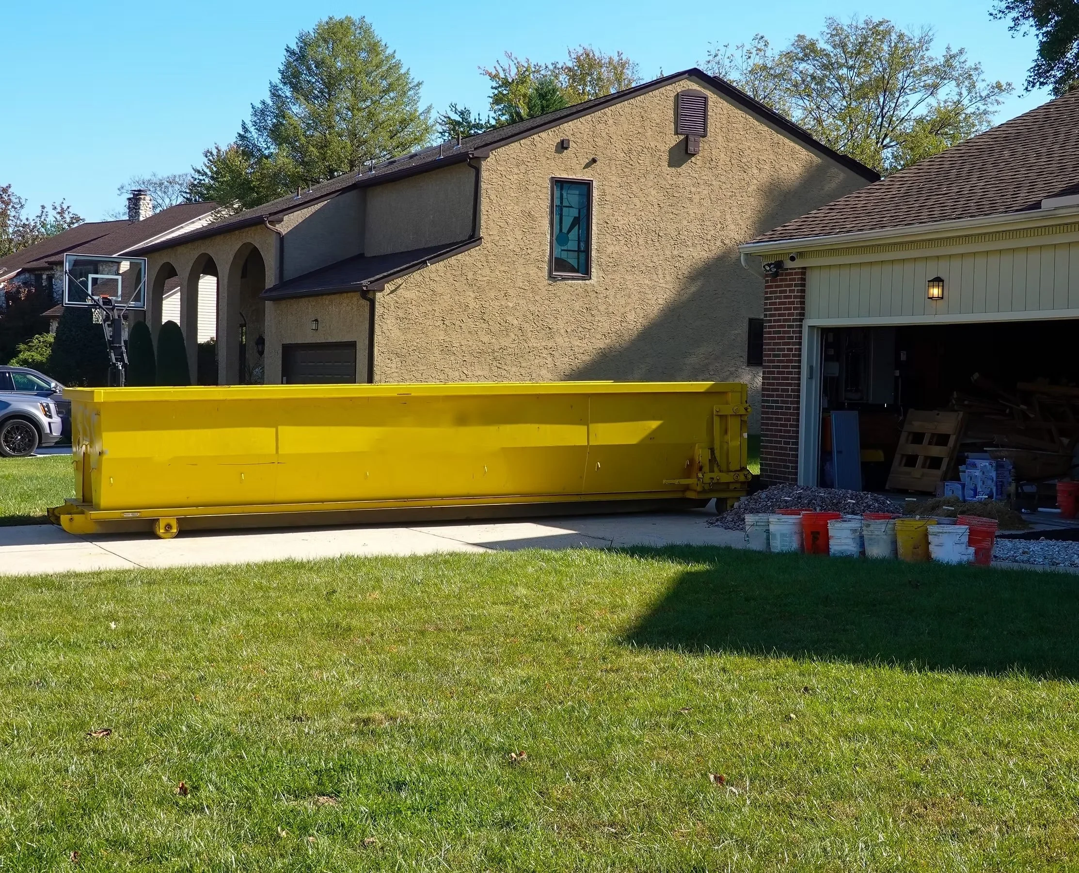 Residential backyard with a yellow construction dumpster in front of a garage, construction materials on the driveway, and a basketball hoop in the background.
