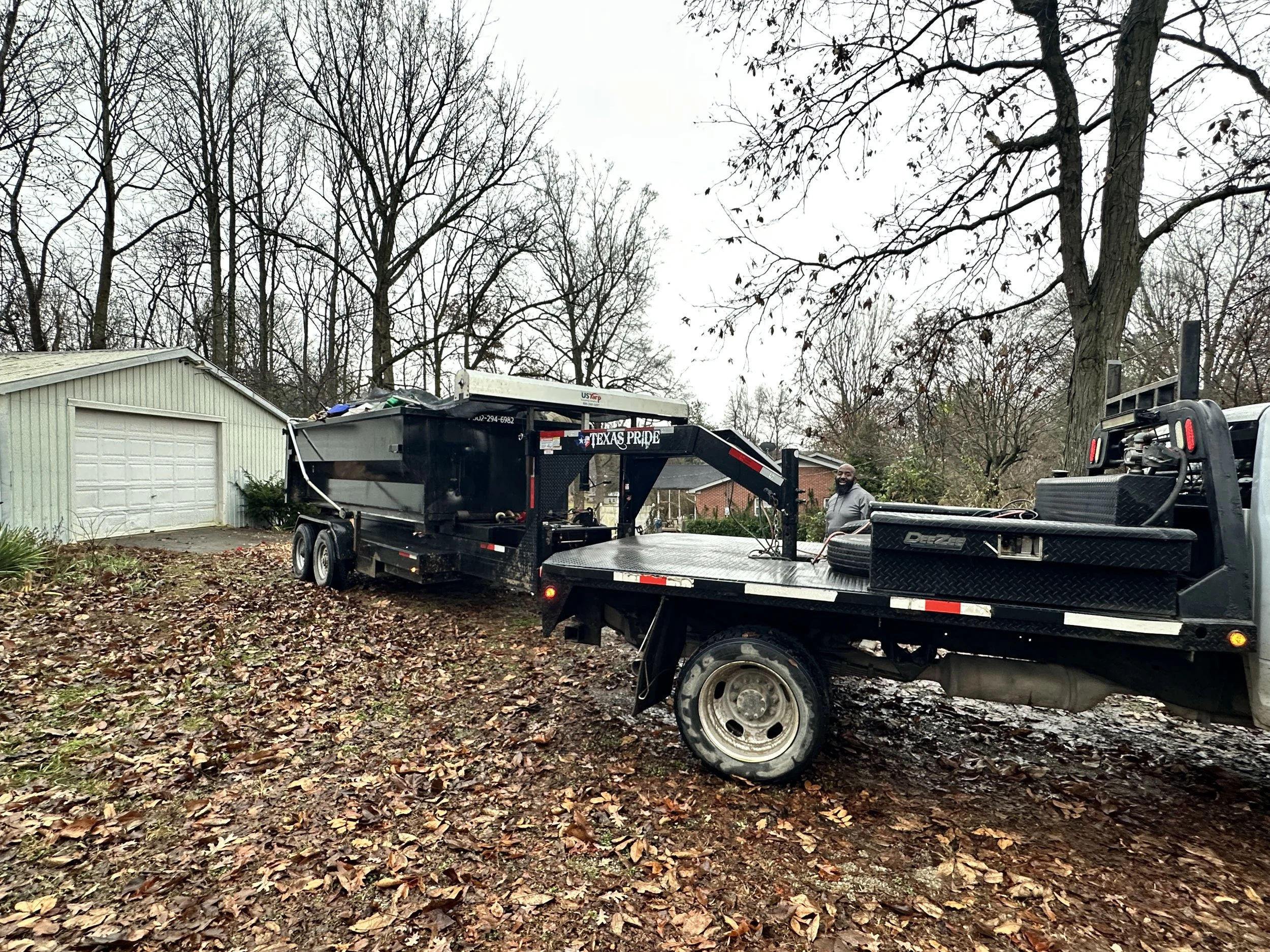 A man standing next to two black utility trailers, one attached to a white truck, parked on a leaf-covered yard with leafless trees and a garage in the background.