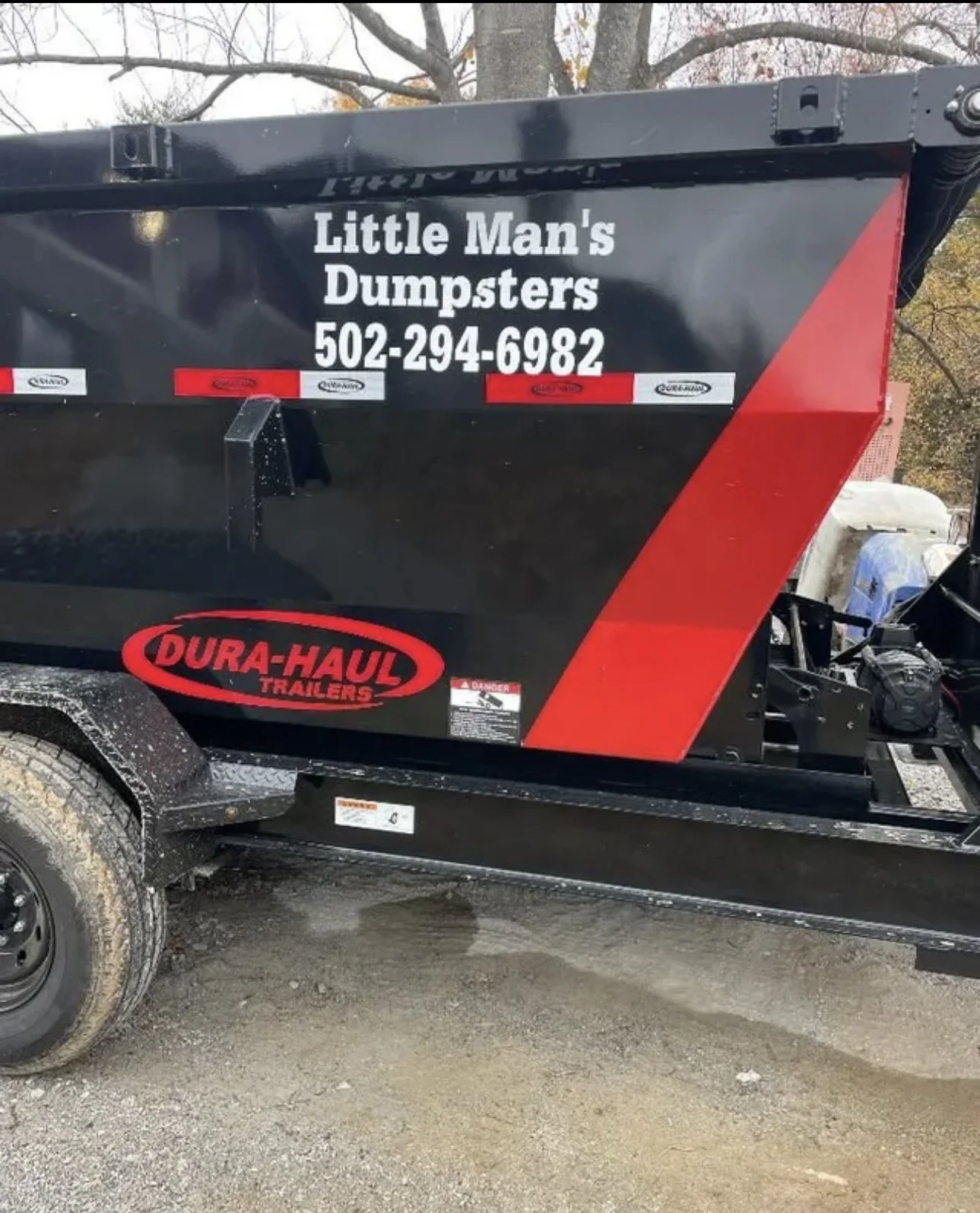 A black and red trailer with a sign that reads 'Little Man's Dumpsters' along with a phone number, parked outdoors on a dirt surface with trees in the background.