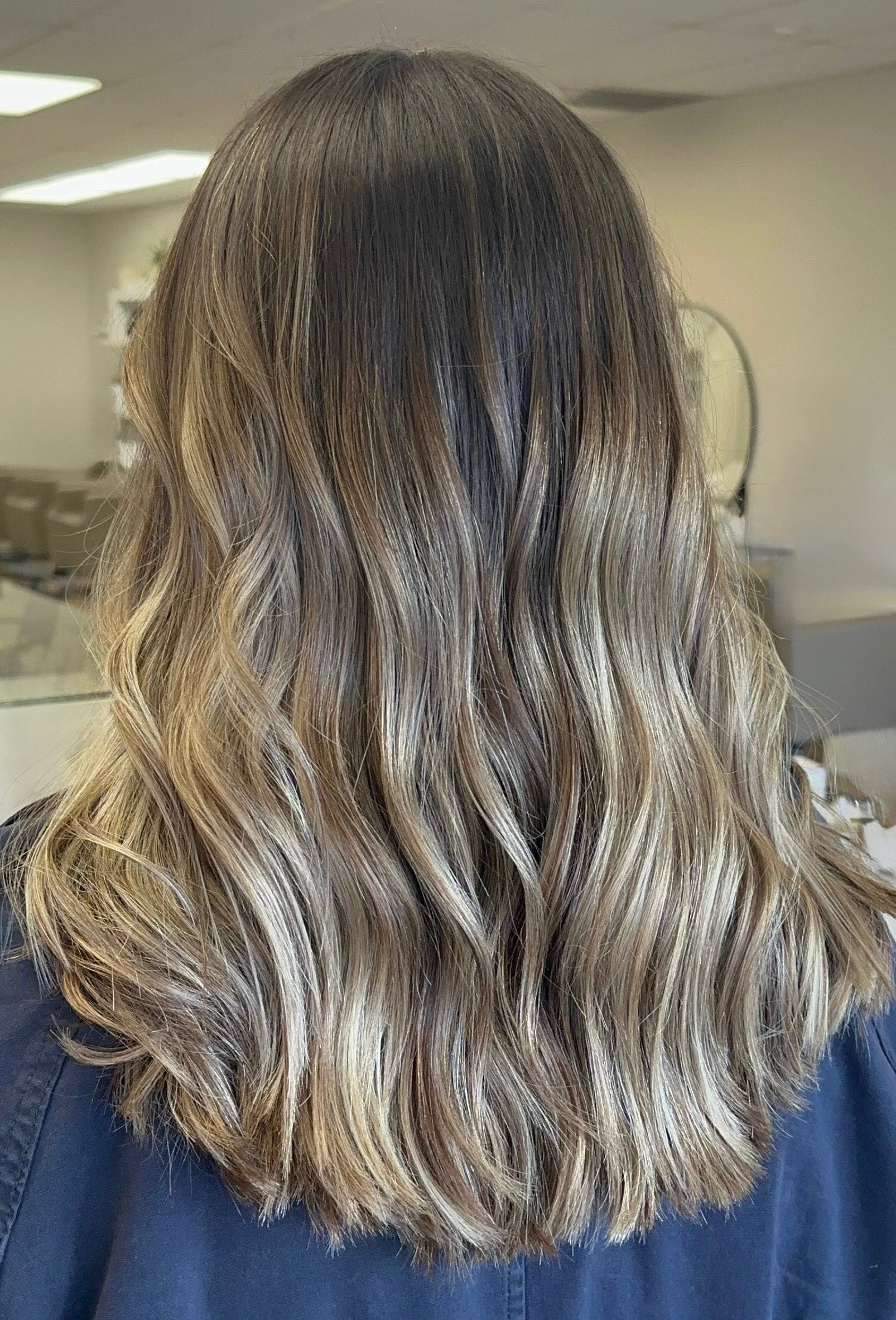 Back view of a woman with shoulder-length, wavy, blonde hair with darker roots, inside a salon.