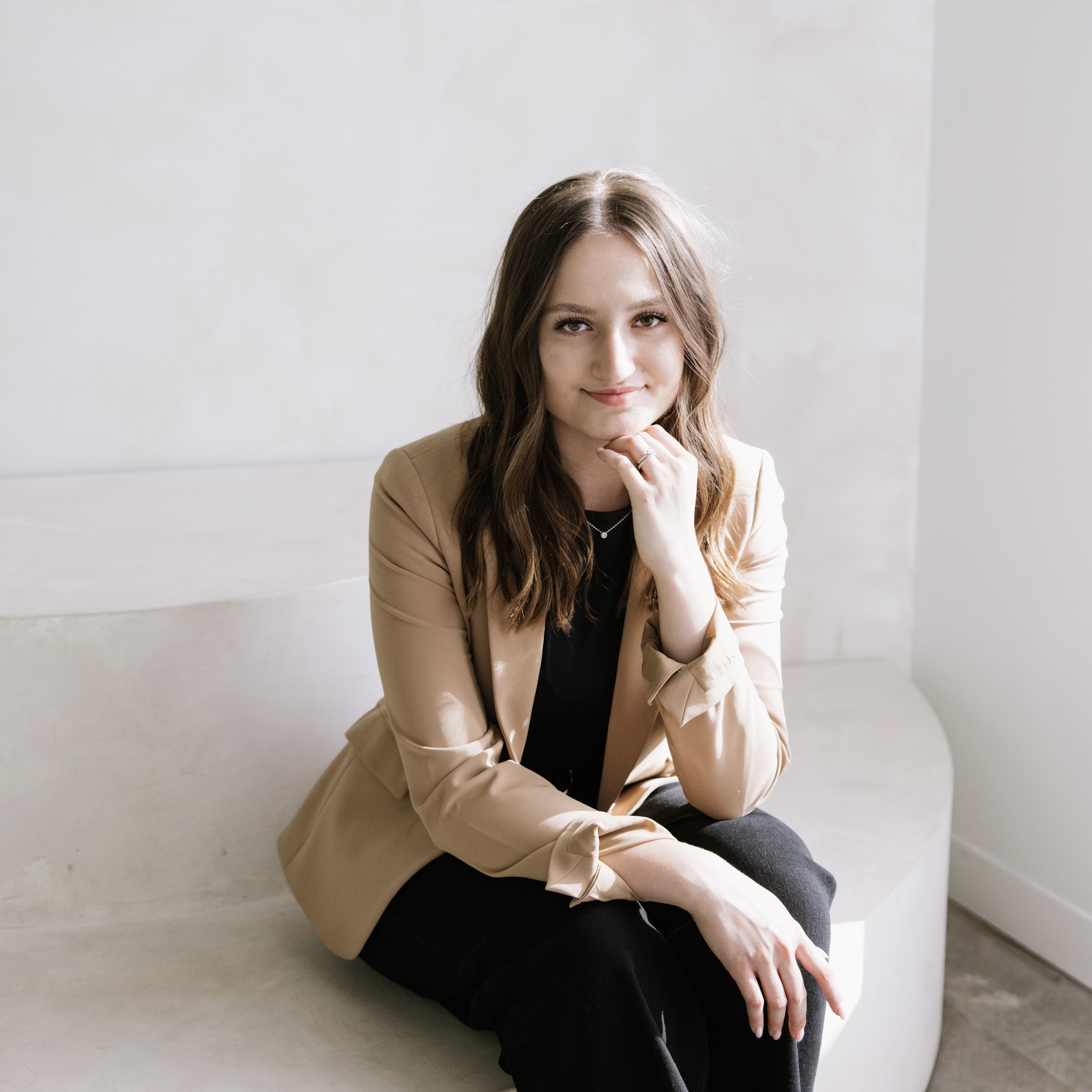 A young woman with wavy light brown hair sitting on a bench, wearing a beige blazer and black clothing, smiling softly and resting her chin on her hand in a bright room.
