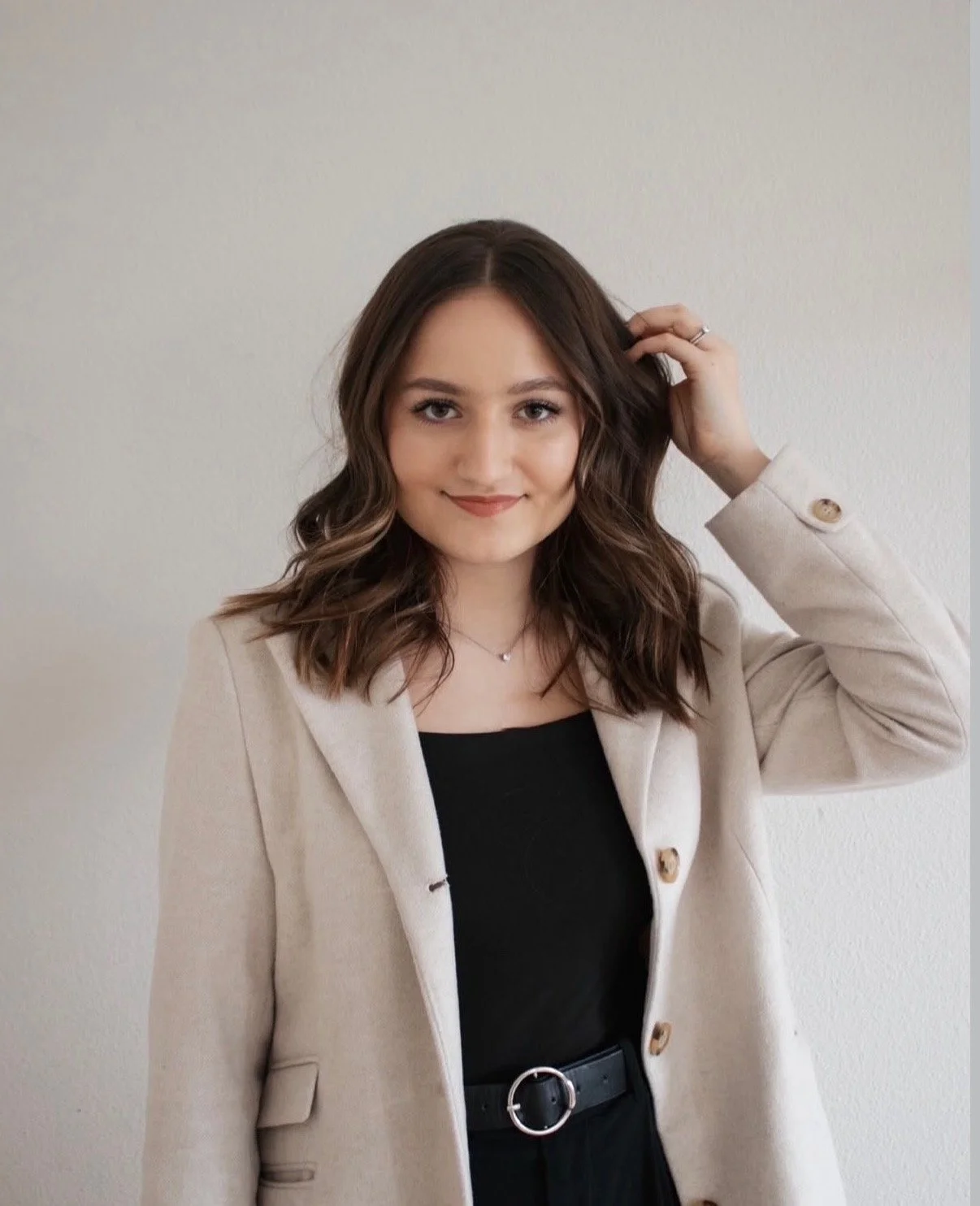 A young woman with shoulder-length brown hair and a slight smile, wearing a beige blazer over a black top, standing against a plain light-colored wall, touching her hair with her right hand.