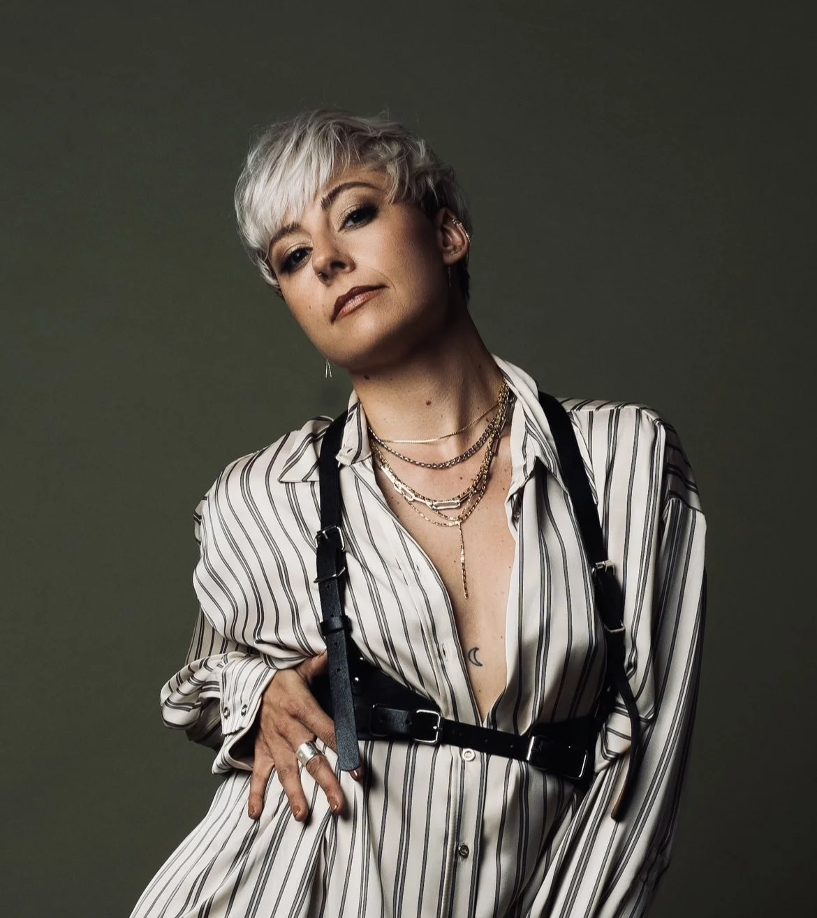 Portrait of a woman with short, platinum blonde hair featuring dark roots, wearing layered necklaces, earrings, a striped shirt with the top buttons undone, and a black strap across her chest, against a dark green background.
