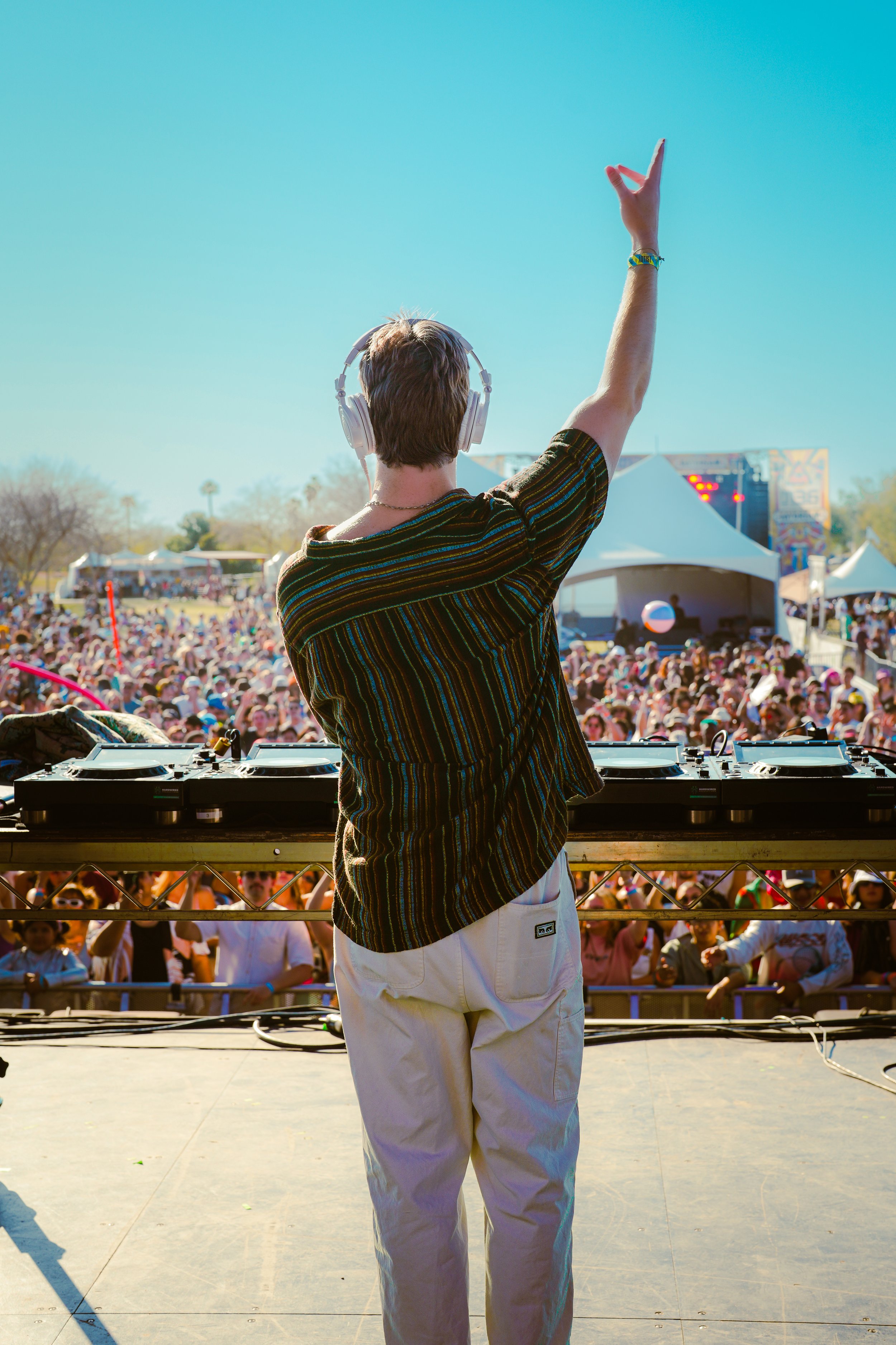 DJ performing on stage at an outdoor music festival with a large audience and a clear blue sky.