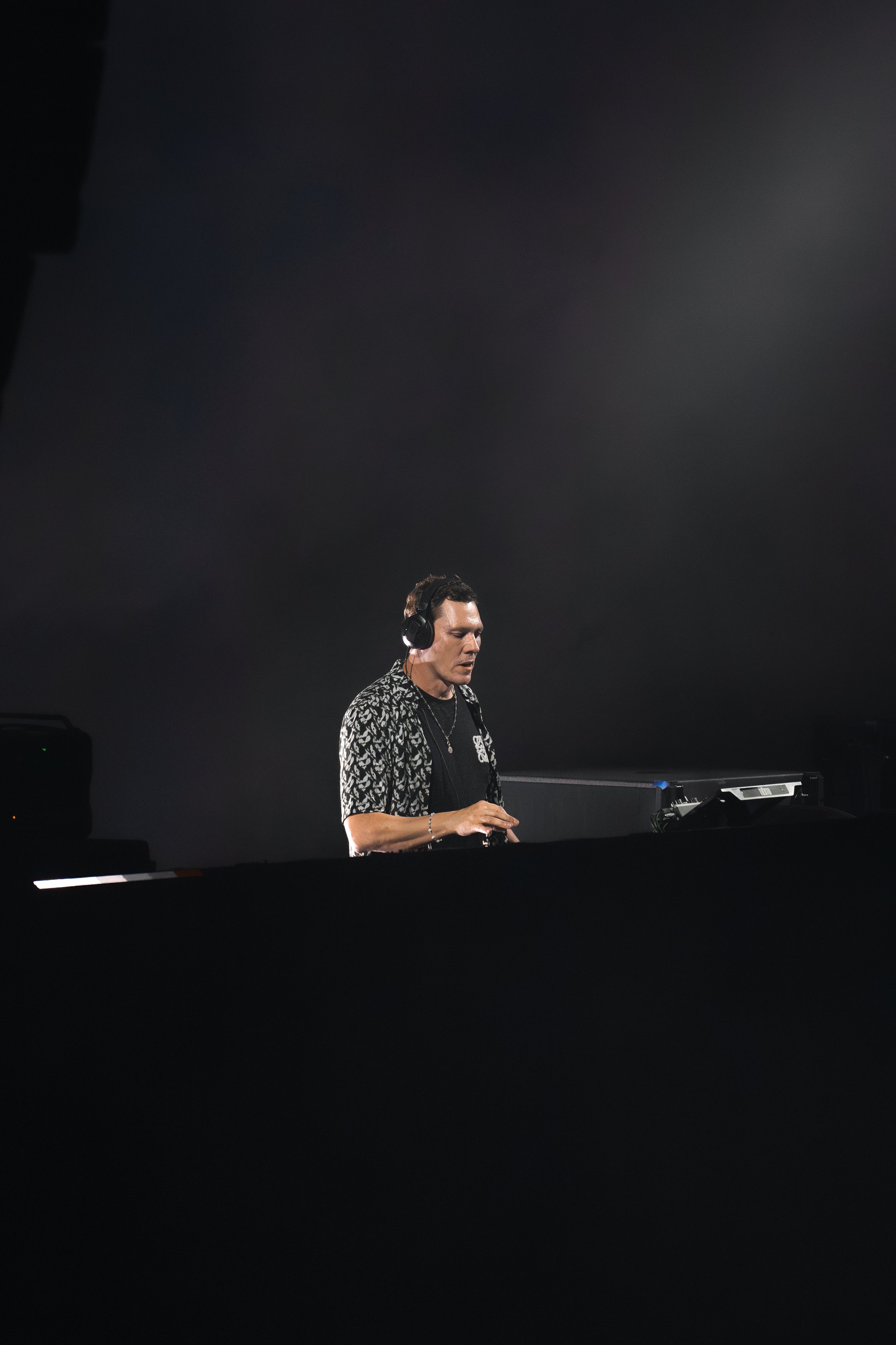 A DJ wearing headphones and a patterned shirt is performing at a dark venue, standing behind a DJ setup with equipment.