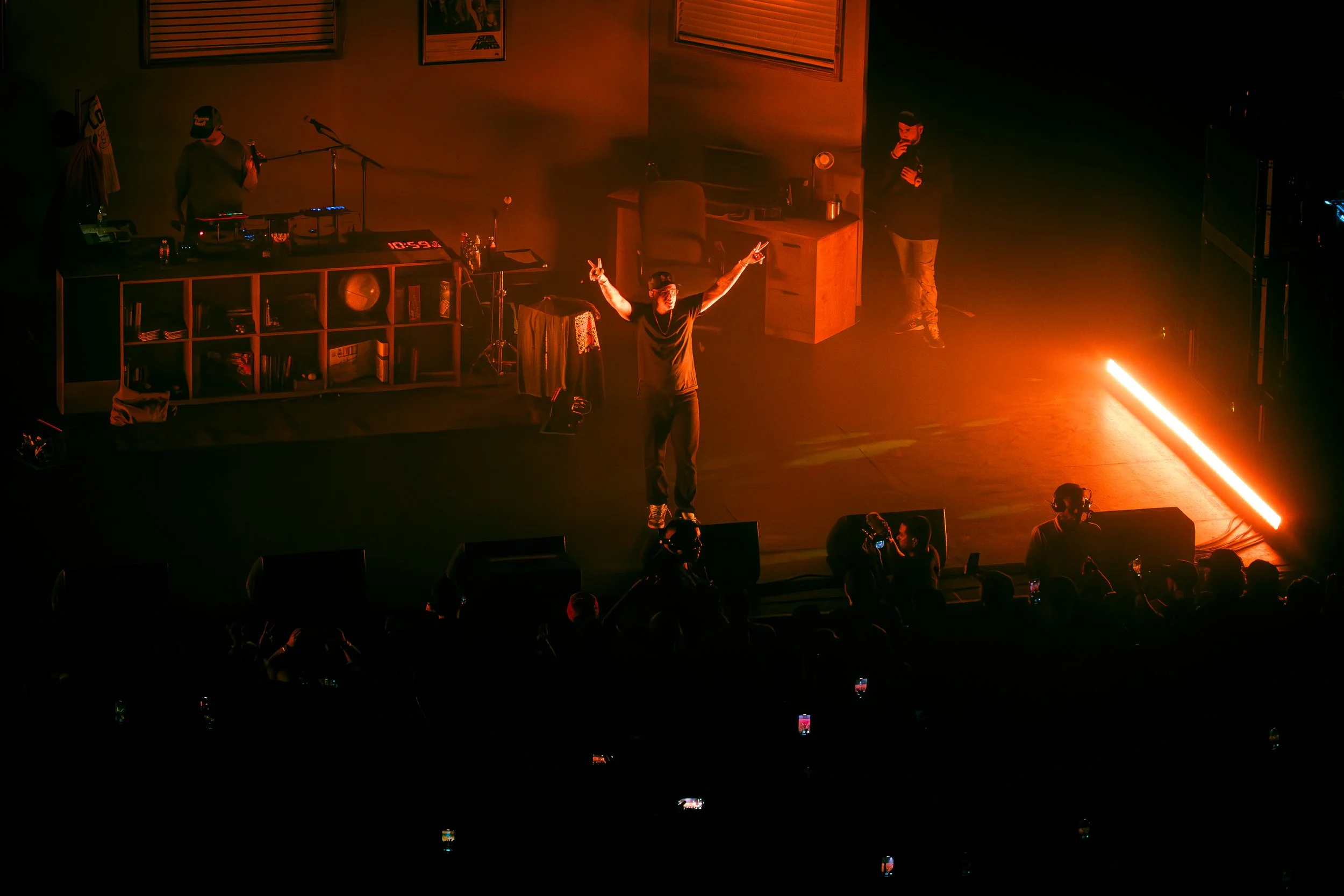 A performer on stage with arms raised making peace signs, illuminated by orange stage lighting, with a DJ and a person standing near a desk in the background, and an audience with phones capturing the event.