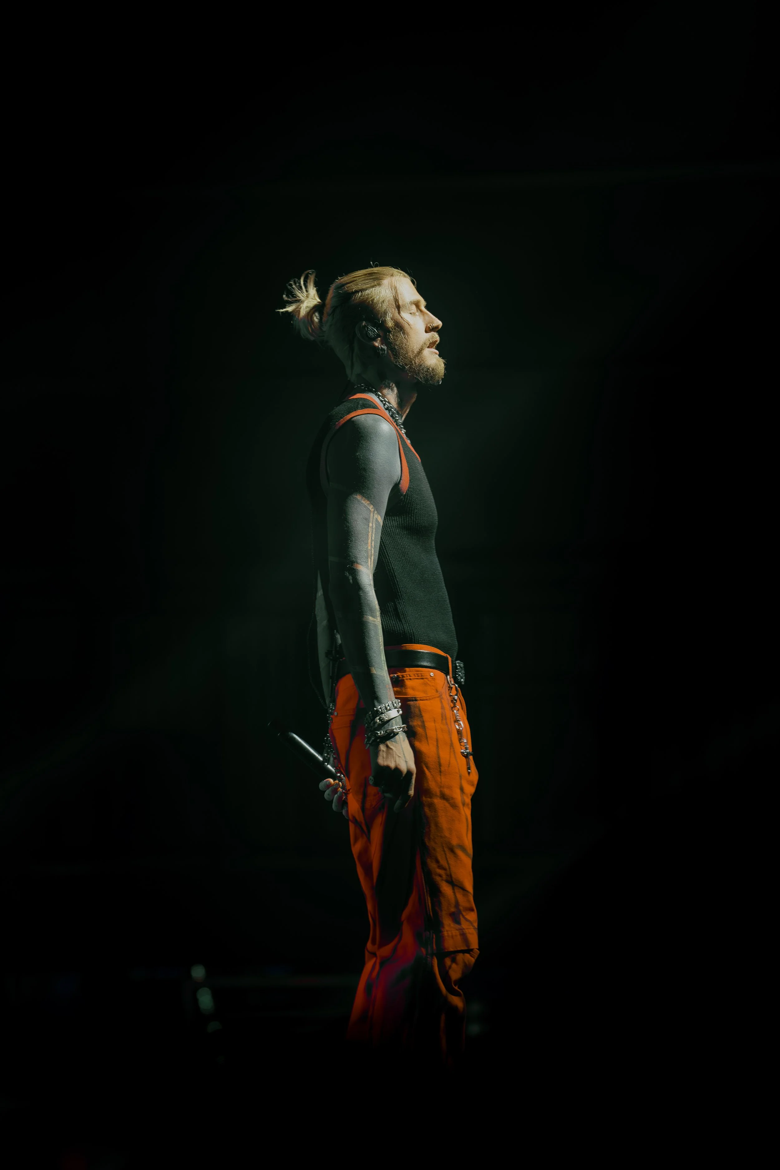 A male performer on stage with eyes closed, wearing an orange and black outfit, holding a microphone, with a spotlight highlighting his profile against a dark background.