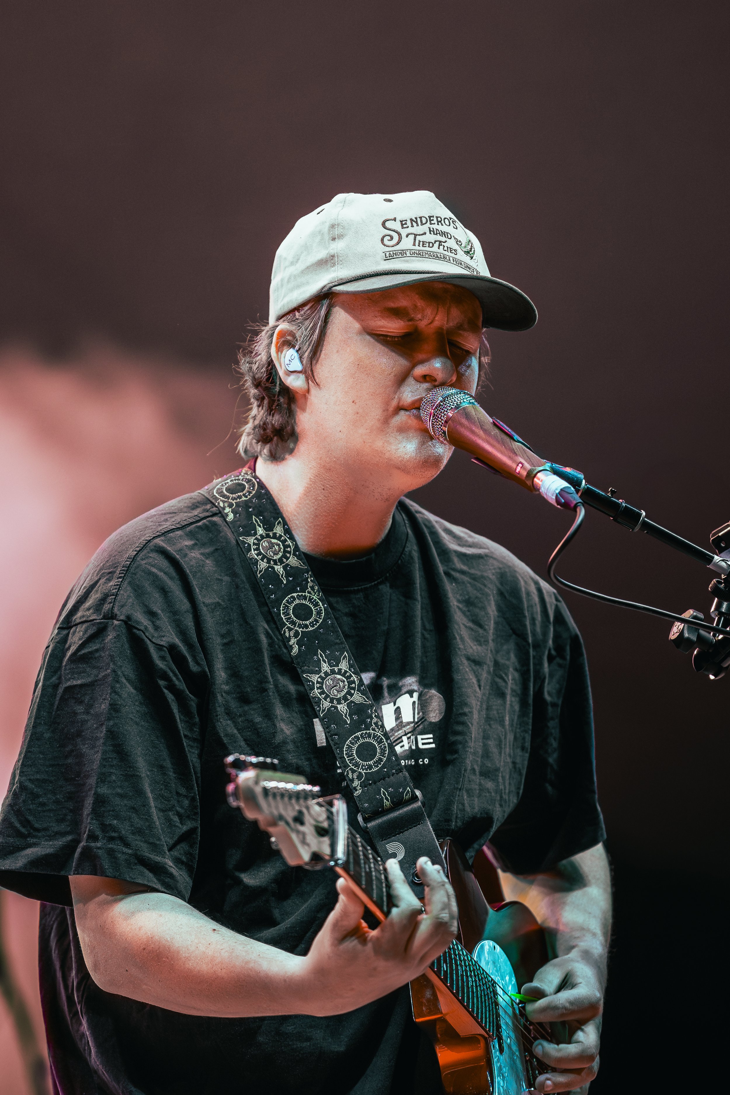A young man performing on stage, singing into a microphone and playing an electric guitar, wearing a gray cap and a black t-shirt with a strap across his chest.