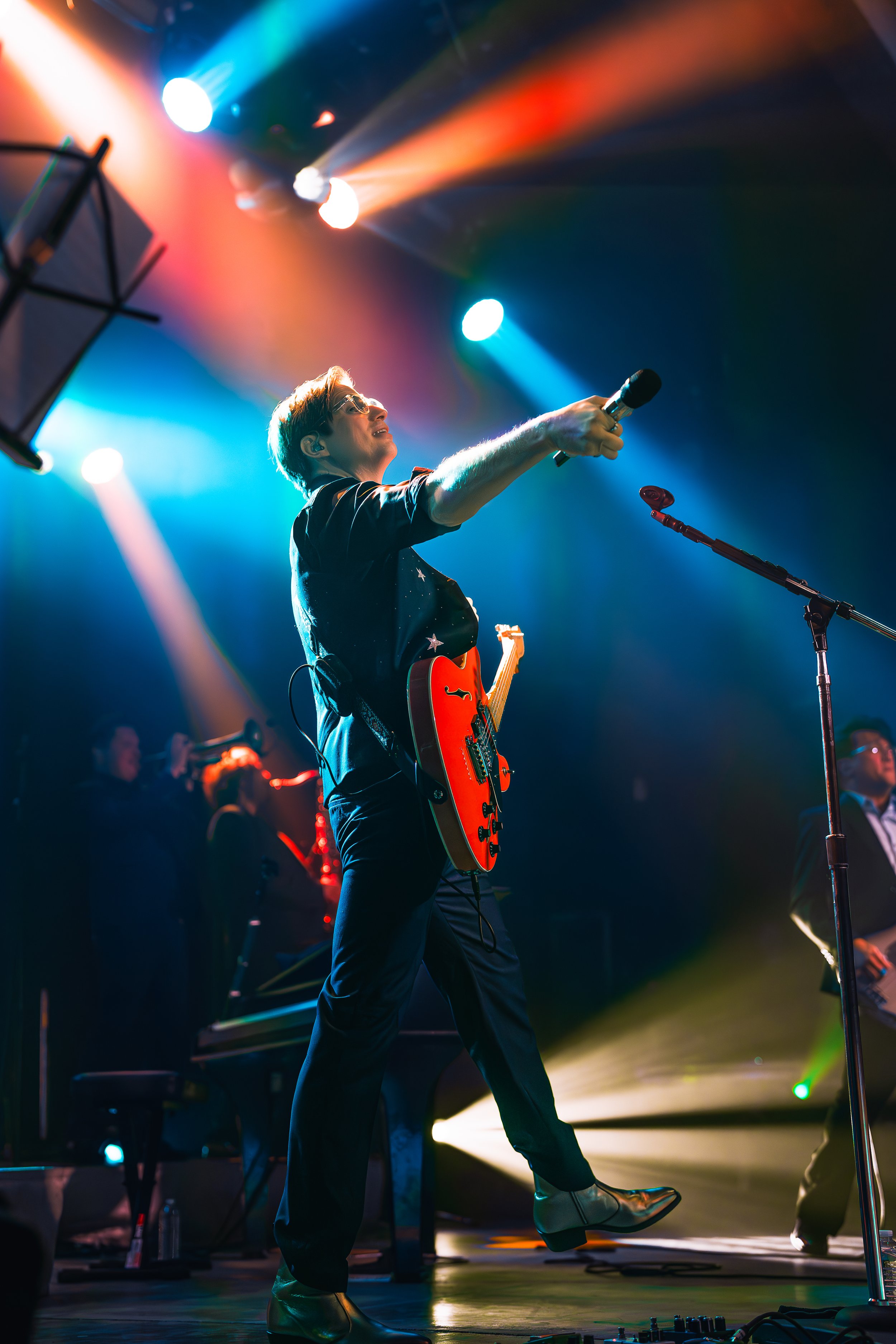 Musician on stage with red electric guitar, raising microphone, colorful stage lights, and other band members in background.