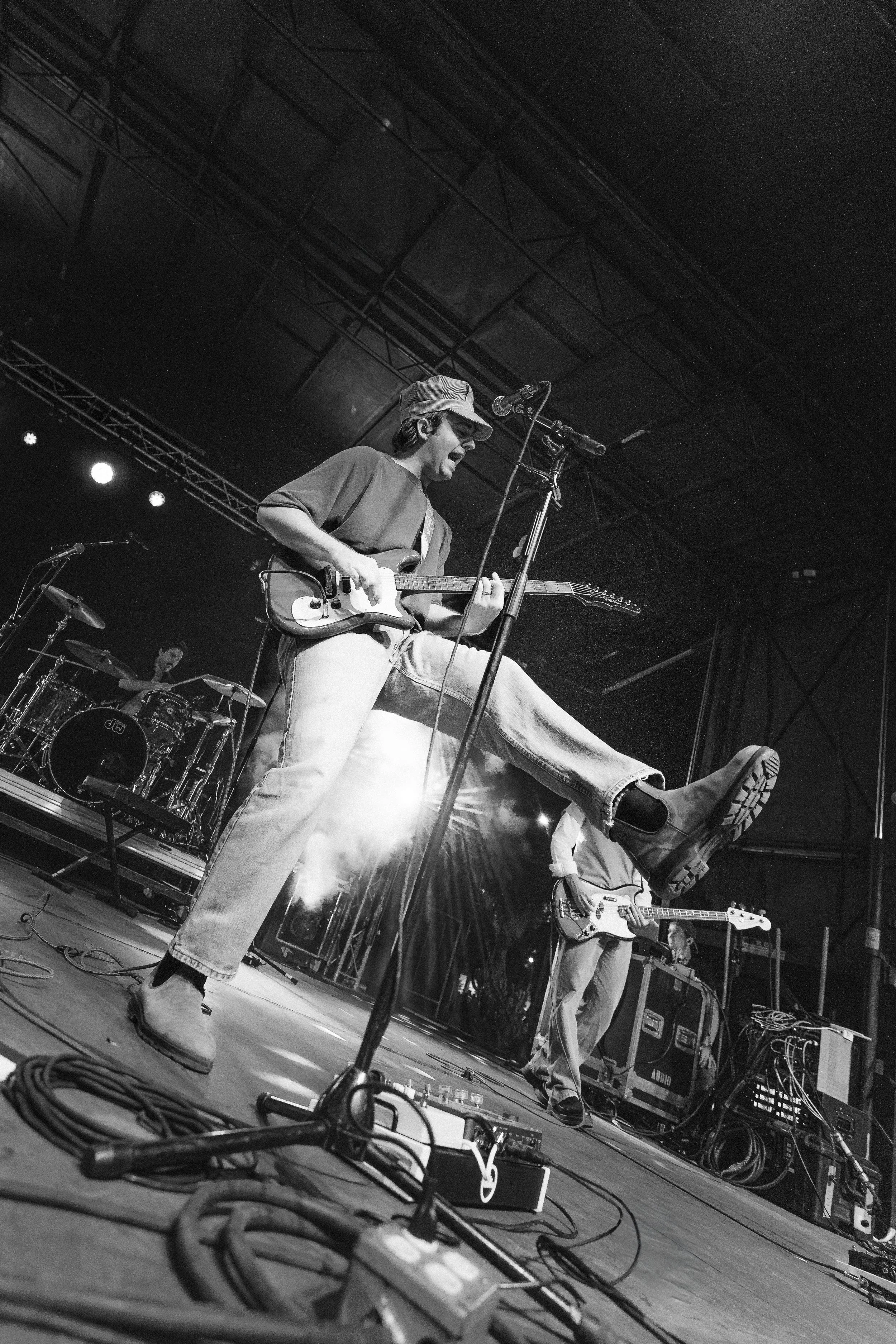 A black and white photo of a band performing on stage, with the lead guitarist mid-air jump, playing an electric guitar. The drummer and another guitarist are visible in the background.