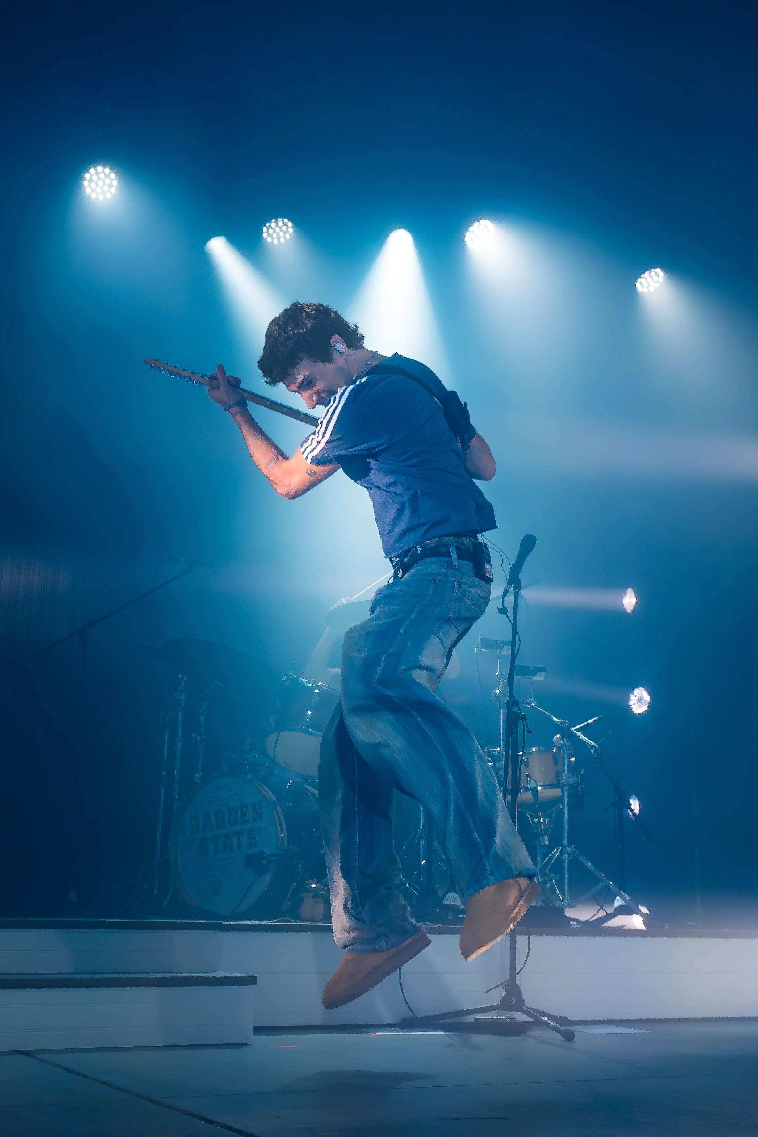 A musician playing an electric guitar on stage, jumping with a microphone stand nearby, with blue stage lights and a drum set in the background.