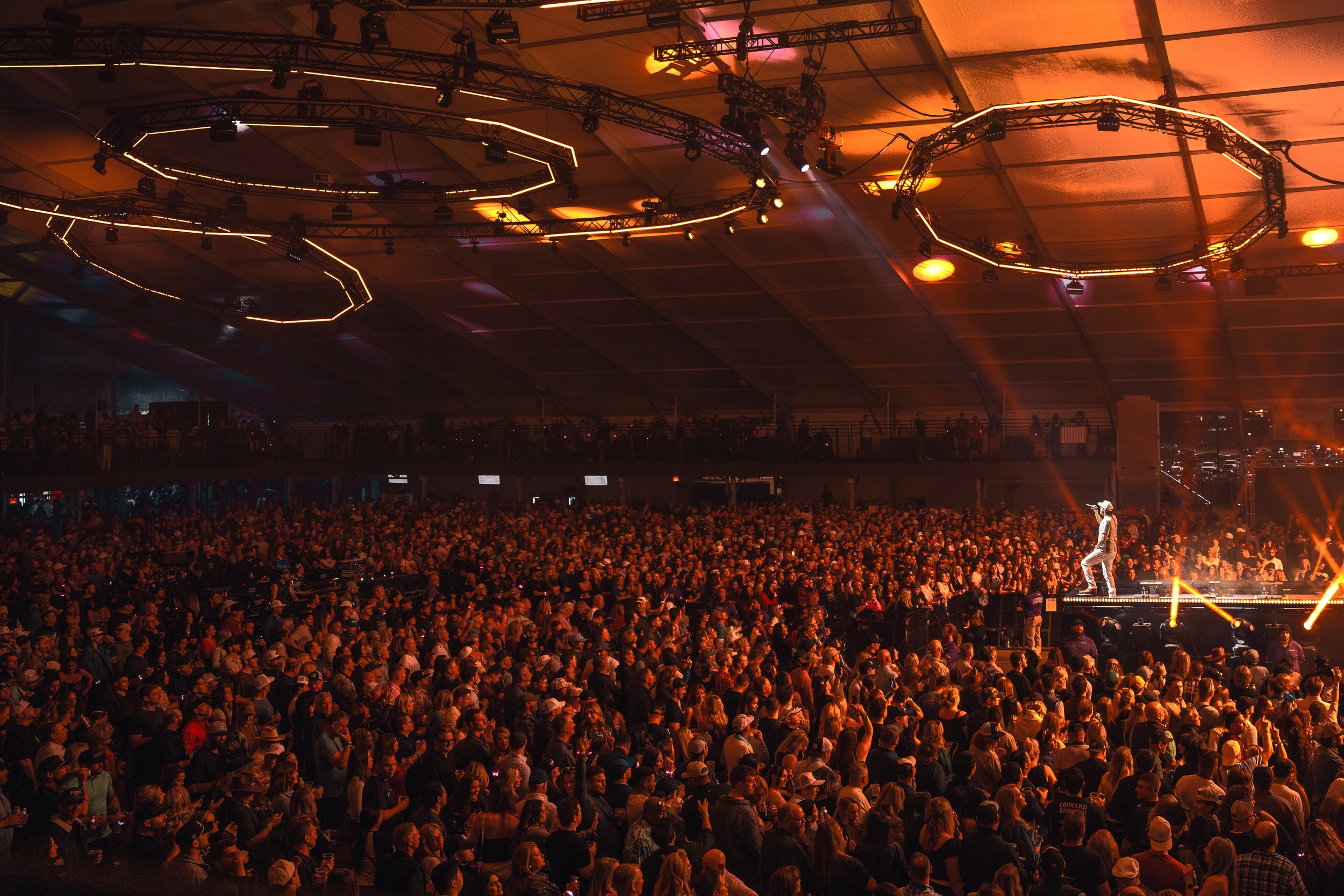 A large indoor concert with a big crowd, stage lights, and a performer on an elevated stage.