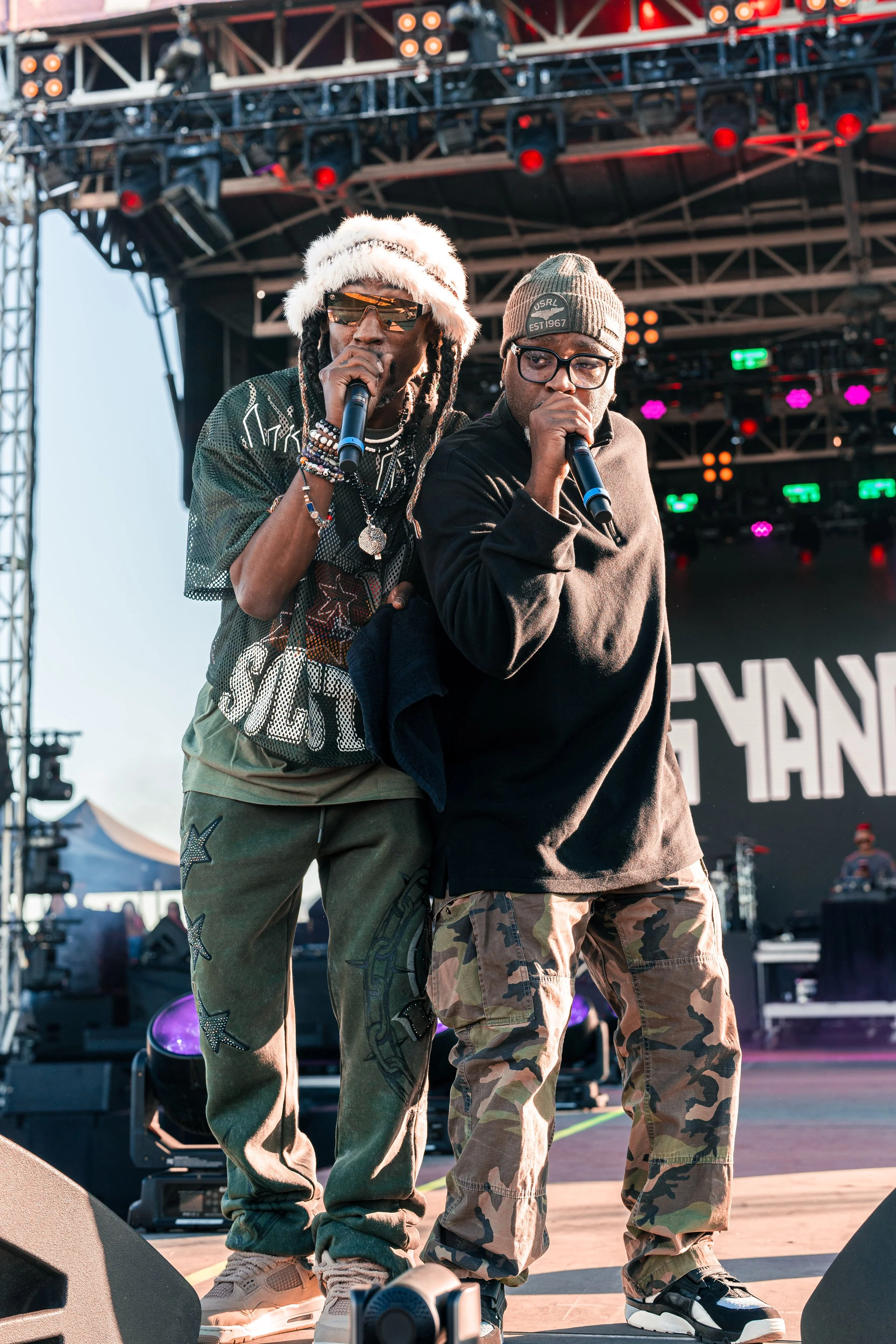 Two male performers on stage holding microphones during a concert, with a large audience and colorful stage lights in the background.