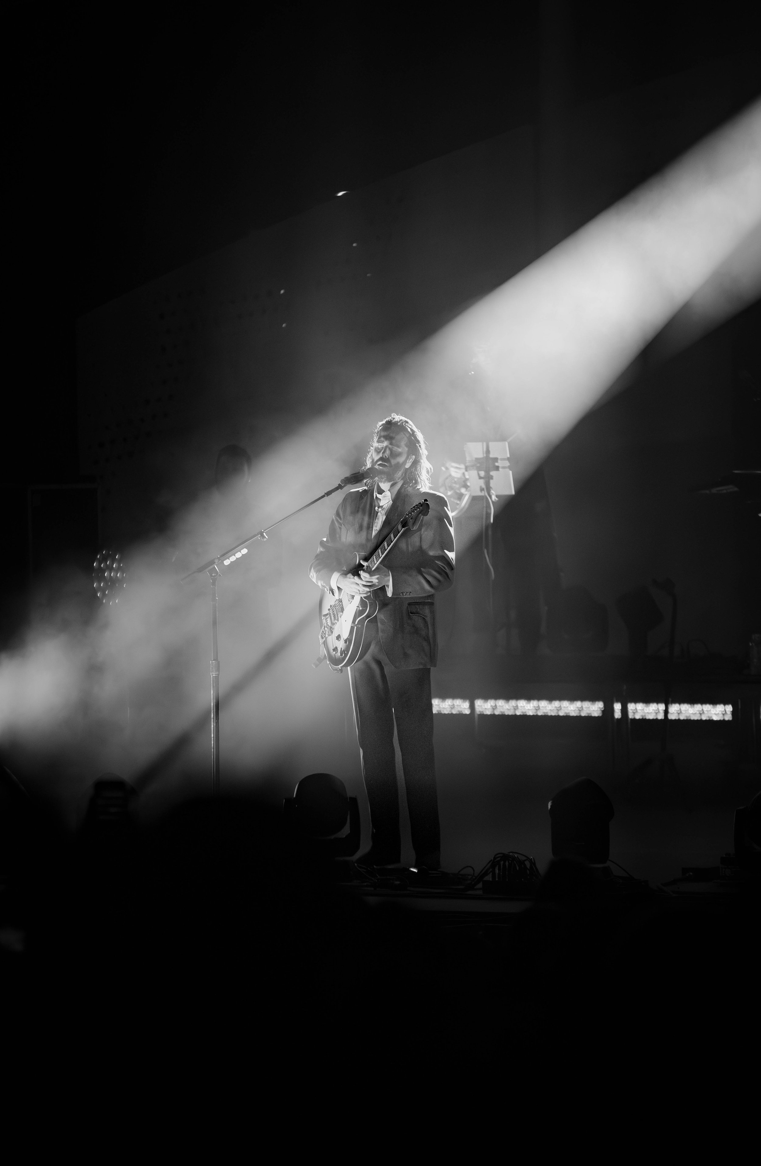 A black-and-white photo of a musician performing on stage with an electric guitar, standing in a spotlight with a microphone in front of him, and fog or smoke around him.