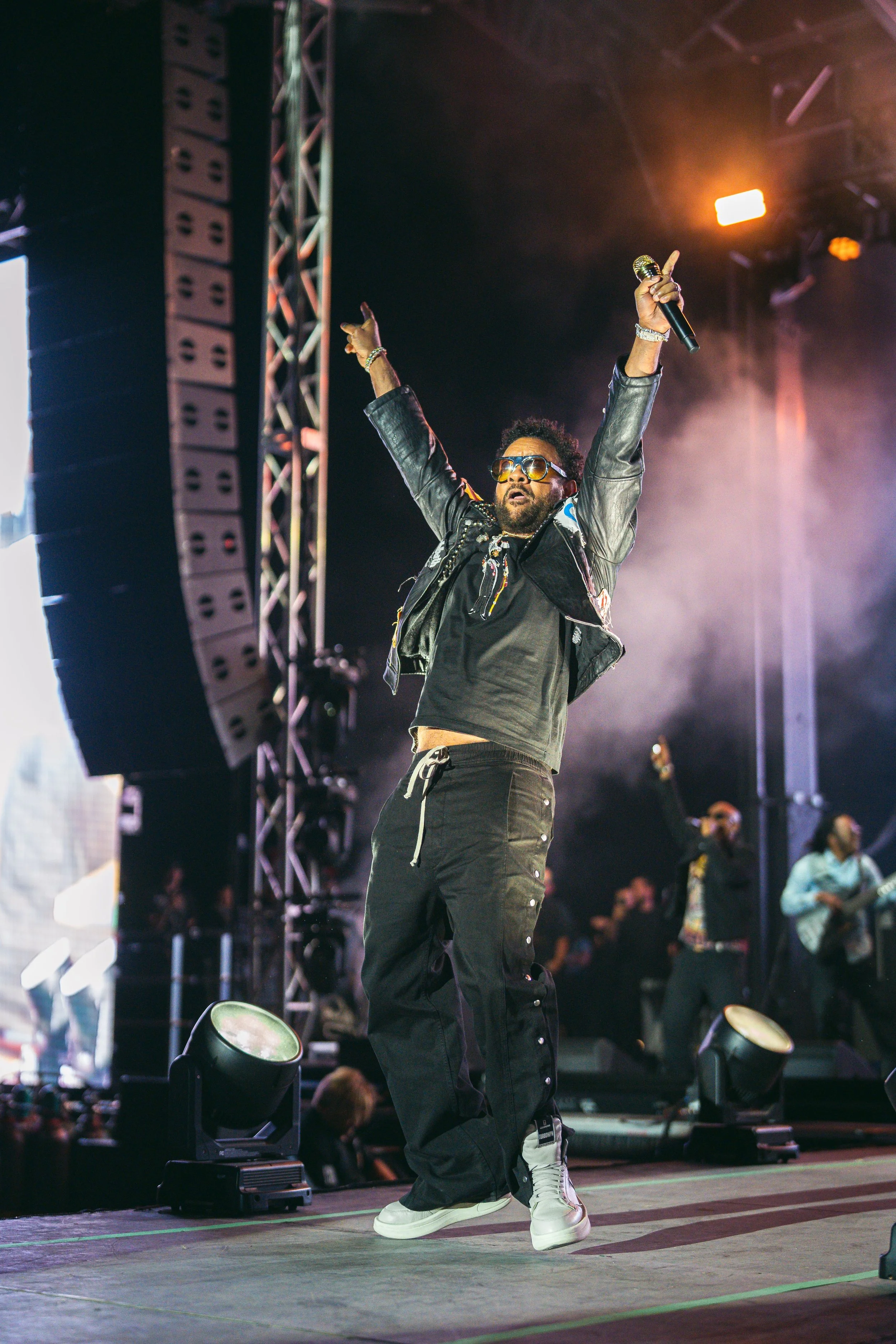 A male performer dancing on stage with arms raised, wearing sunglasses, a black jacket, black pants, and white sneakers, under stage lights with a microphone in hand during a concert.