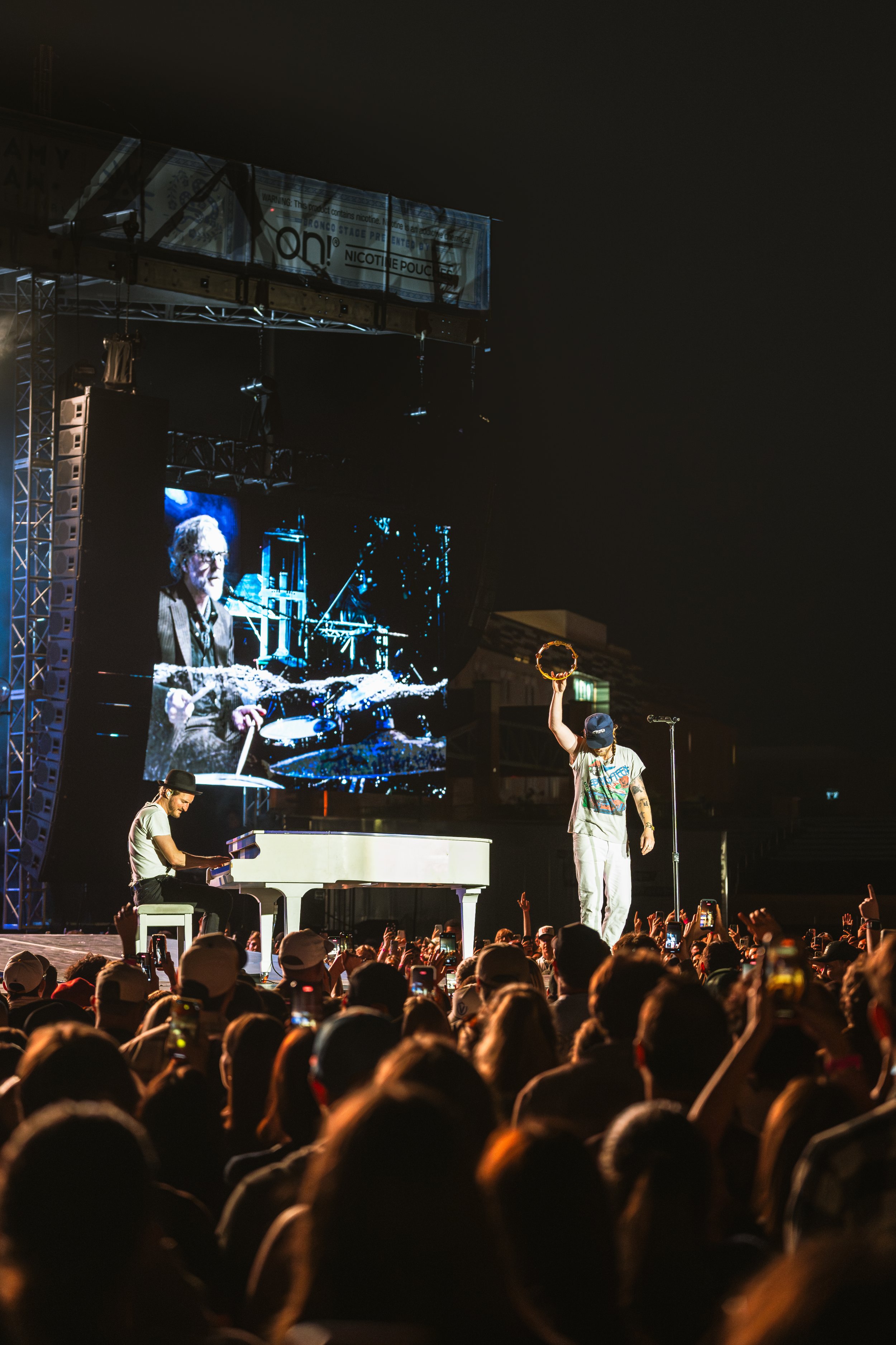 A concert scene at night with a singer holding a tambourine on stage and a pianist playing a grand piano. A large crowd is in front of the stage, many holding up phones to record. A big screen behind shows a close-up of the drummer.