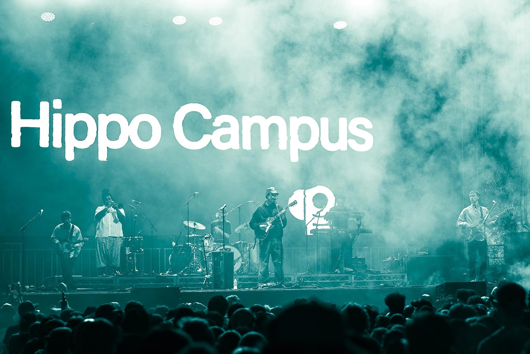 A stage with a band performing live in front of a large crowd, featuring the text 'Hippo Campus' projected on the background.