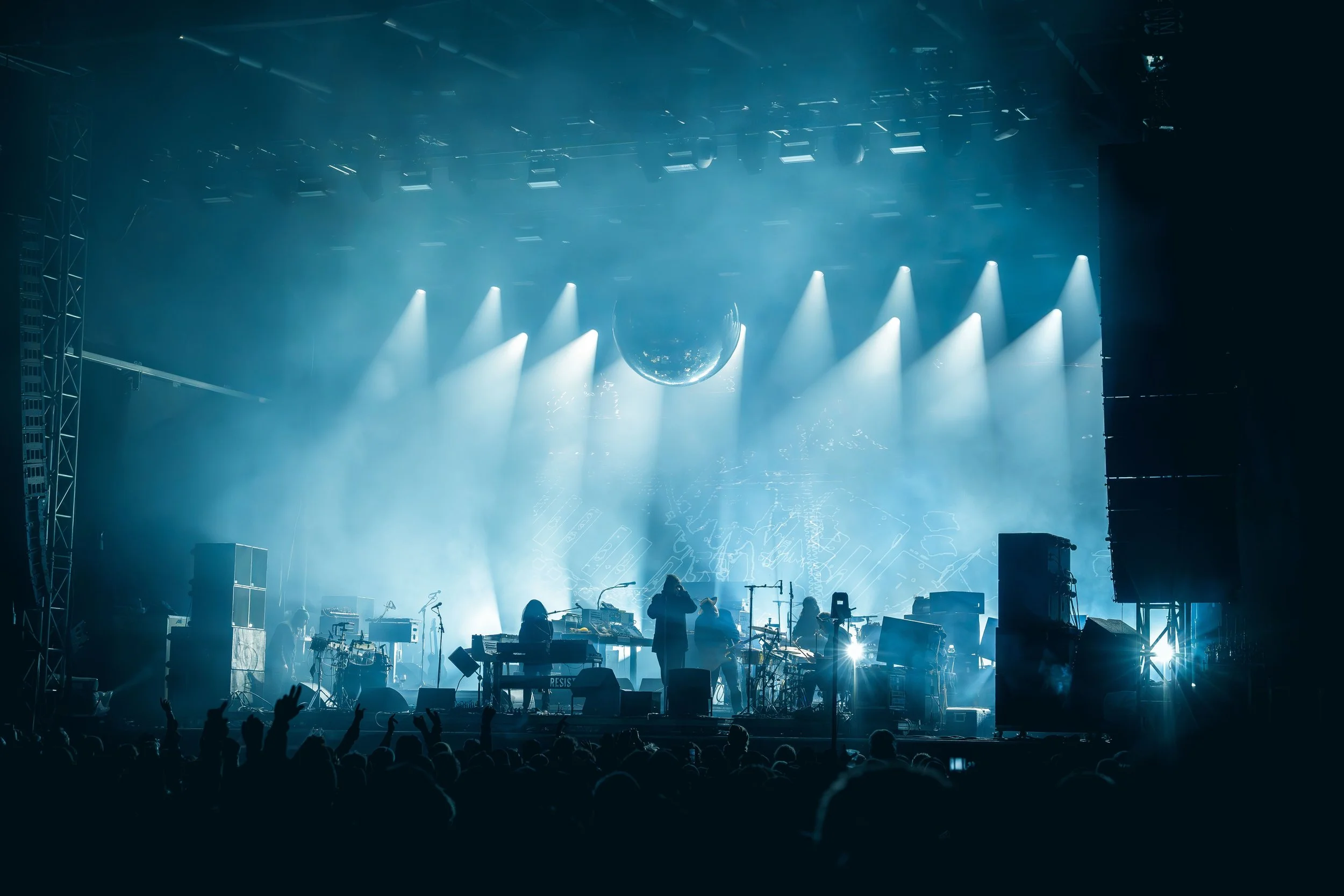 A concert stage lit with blue lights, with silhouettes of musicians and equipment, and an audience in the foreground.