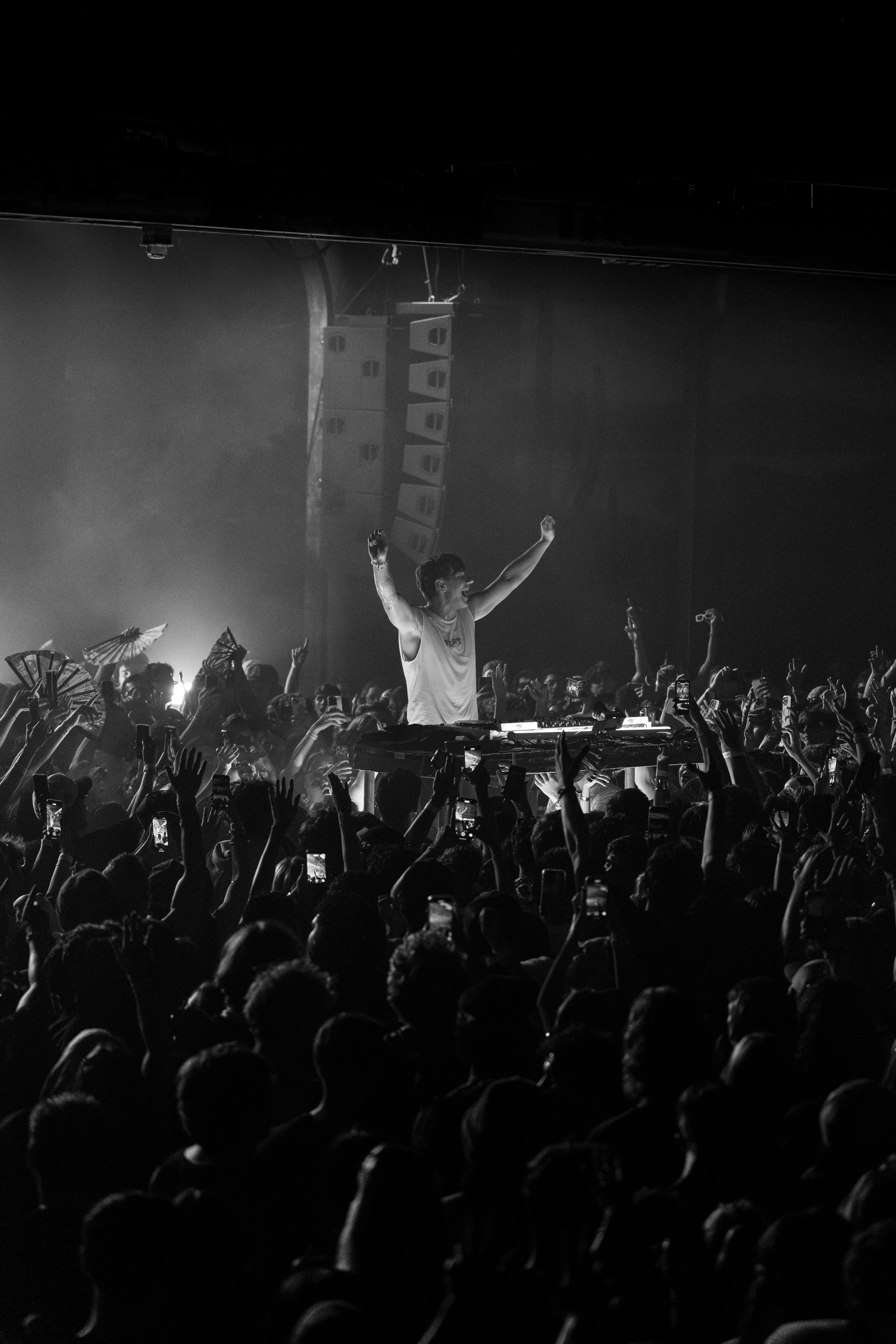 Black and white photo of a DJ performing on stage at a concert with a large crowd dancing and taking photos. The DJ is raising both arms, facing the audience, with sound speakers and lighting in the background.
