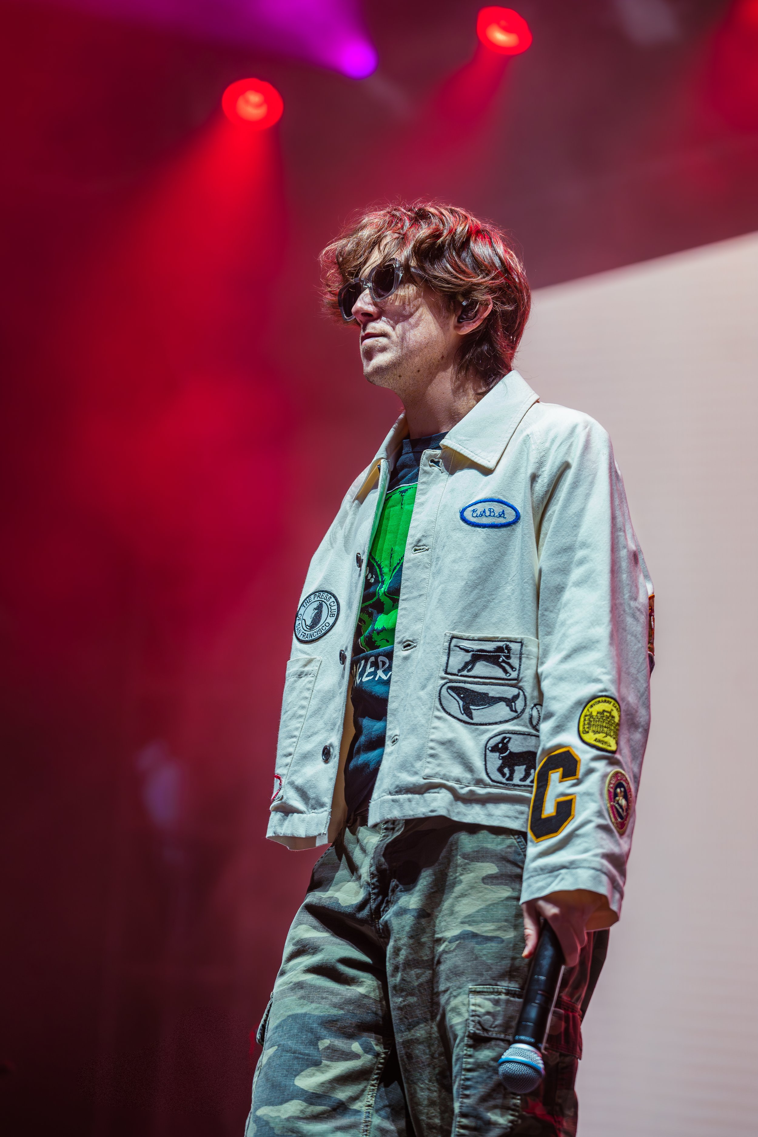 A man with wavy hair, wearing sunglasses, a beige jacket with patches, and camouflage pants, standing on stage in front of colored stage lights, holding a microphone.