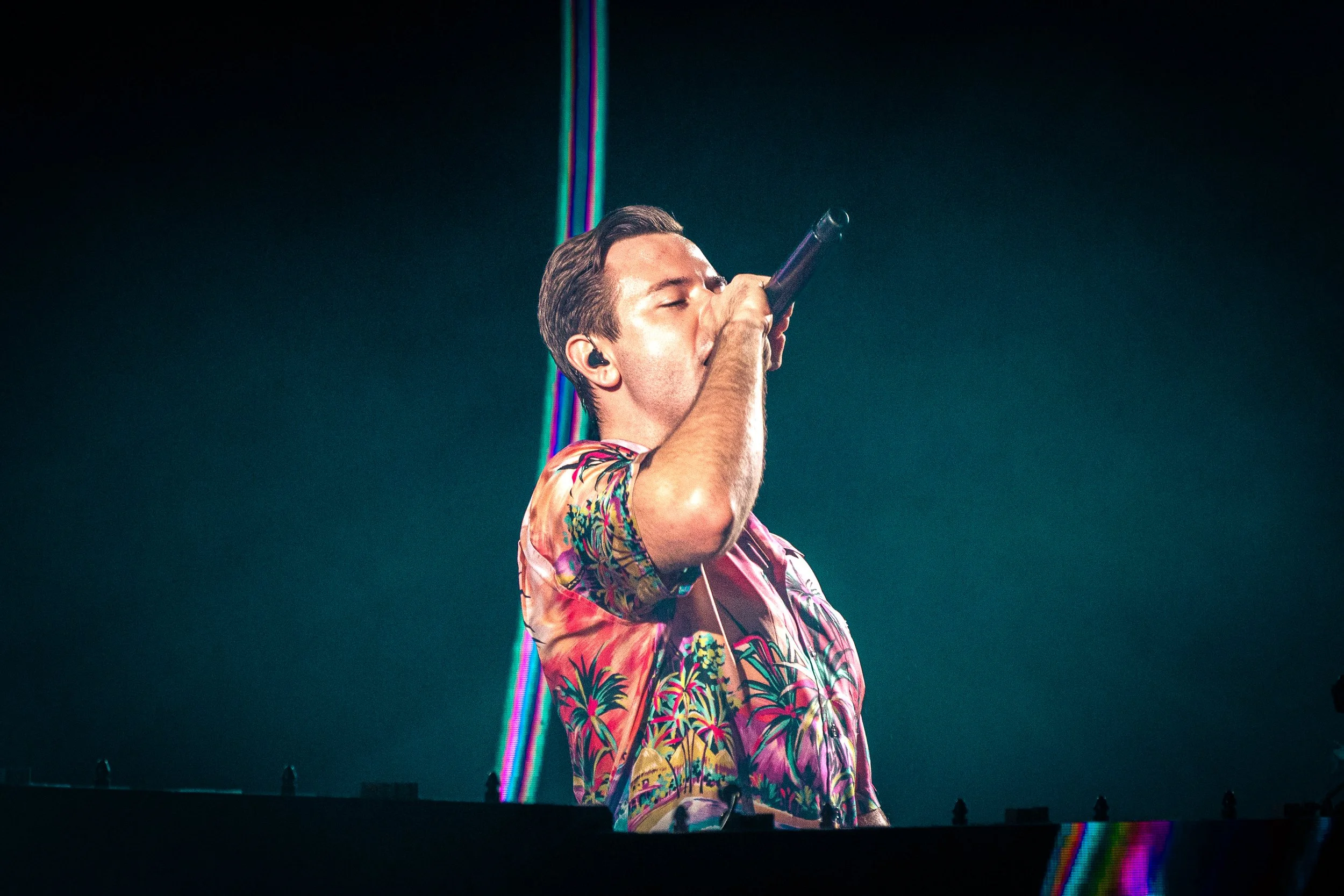 Young male DJ with brown hair wearing a colorful tropical shirt, singing into a microphone during a music performance, illuminated by stage lights.
