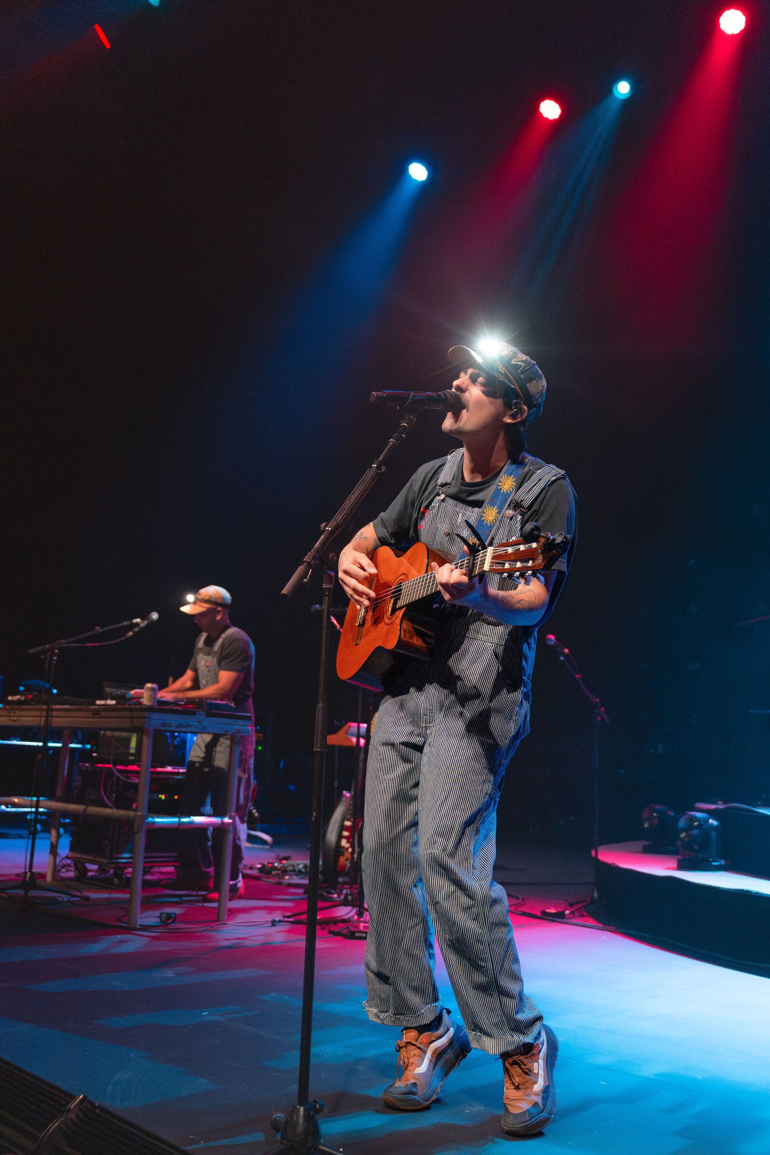 A musician on stage playing an acoustic guitar and singing into a microphone, illuminated by colorful stage lights, with another musician in the background playing a keyboard.