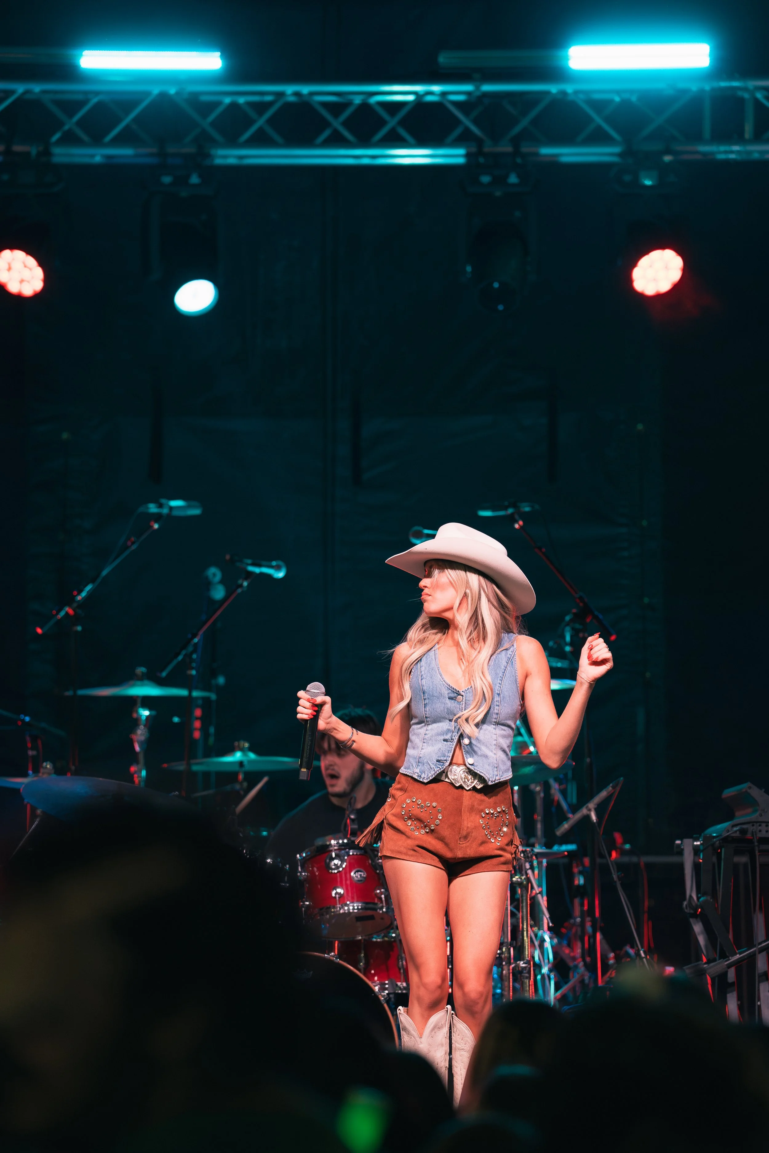 A female country singer performing on stage in front of a microphone, wearing a cowboy hat, denim vest, and brown shorts, with a drummer in the background.