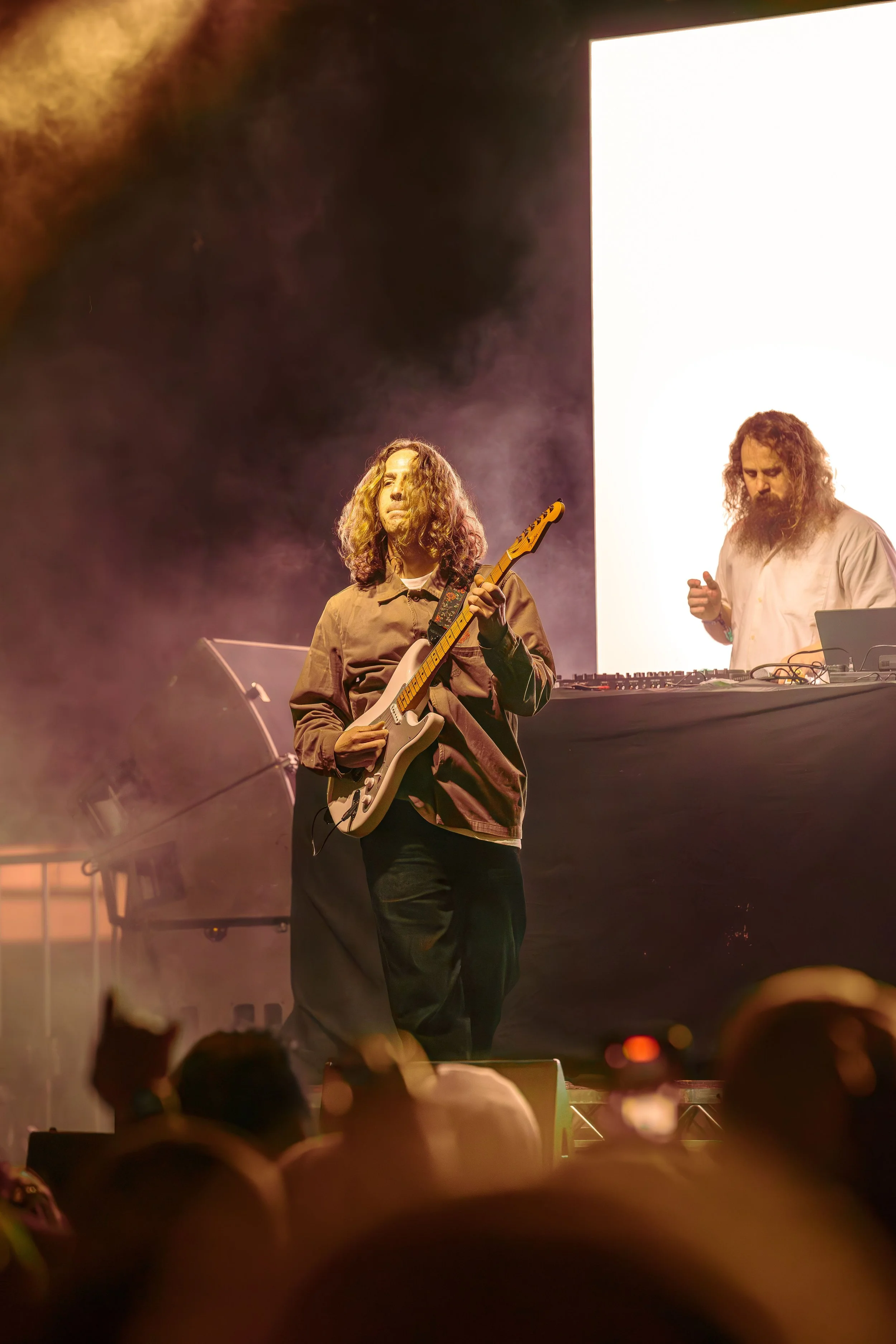 Musicians performing on stage, with one playing an electric guitar and another DJing behind a table with electronic equipment, under stage lighting.