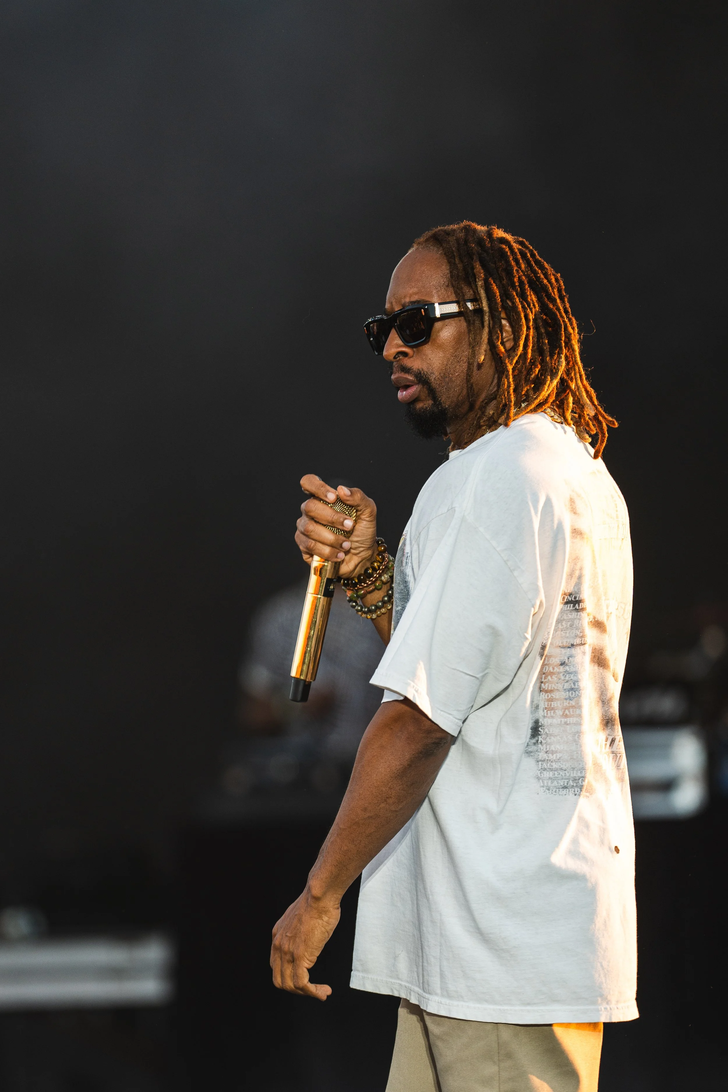 A man with dreadlocks wearing black sunglasses, a white t-shirt, and beige pants is holding a gold microphone during a performance.